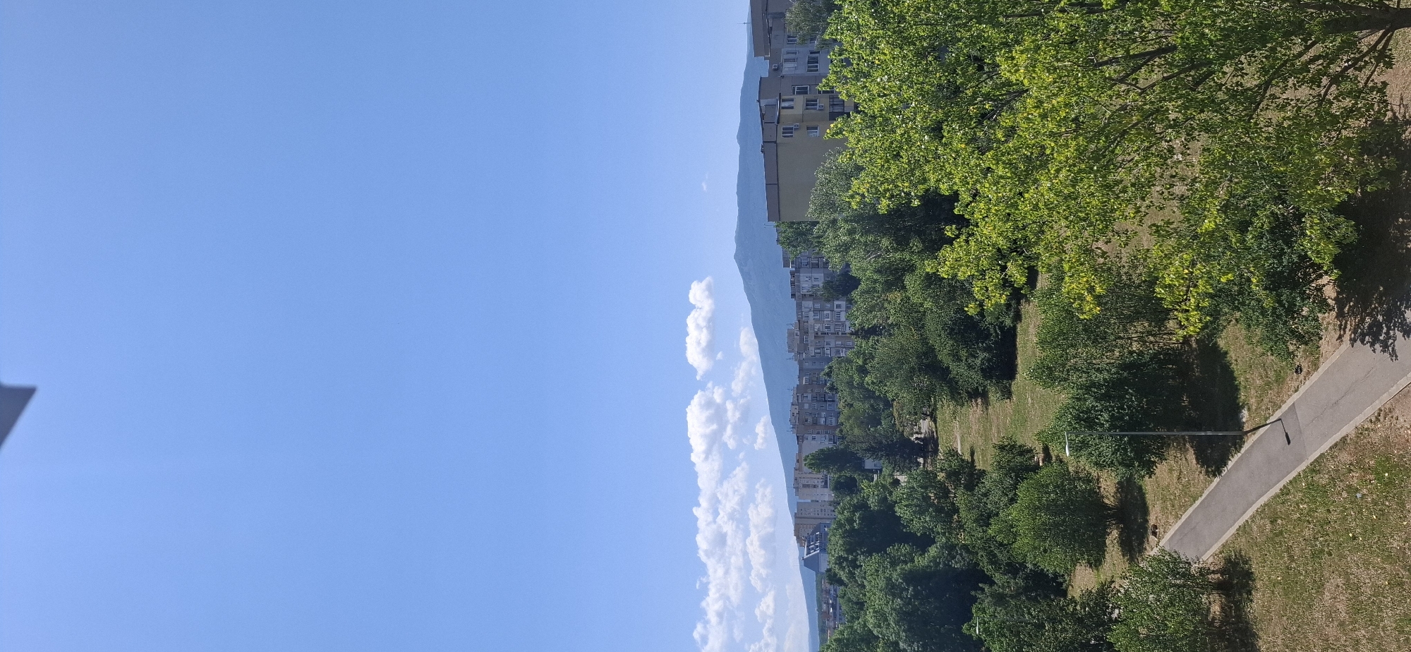 The image is a vertical, wide shot of a cityscape with a mountain range in the background, under a clear blue sky with a few scattered clouds. In the foreground, there is a paved pathway running diagonally from the bottom left towards the middle of the frame, bordered by grass and trees. A tall, slender lamppost is visible next to the pathway.
The middle ground is dominated by a dense array of green trees of various sizes, creating a park-like atmosphere. Beyond the trees, a row of apartment buildings of varying heights and architectural styles stretches across the frame. The buildings appear to be residential, with visible windows and balconies. Some of the buildings are taller and more modern, while others are older, block-style structures.
In the background, a large, imposing mountain range rises against the horizon. The mountain slopes are covered in a muted green or possibly purple hue, indicating distance and vegetation. The overall impression is one of a city integrated with nature, with green spaces and mountains forming a backdrop to the urban environment.
While the specific location within Sofia, Bulgaria is not explicitly indicated by any text or landmarks, the presence of the mountain range suggests a view towards Vitosha Mountain, which is a prominent natural landmark to the south of Sofia. The architecture of the apartment buildings is also consistent with typical residential construction in Bulgarian cities. There are no people or specific actions depicted in the image.