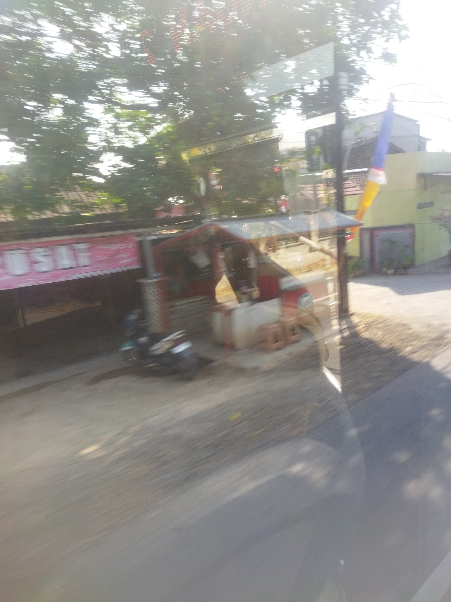 The image shows a blurred scene of a street in Nambangan, Indonesia, captured from a moving vehicle. In the foreground, a road surface is visible, with some debris and shadows. To the left, a building with a pink banner displaying white text that appears to read "PUSAT" is partially visible. Adjacent to this is a small stall or kiosk with a red roof and various objects inside, including what looks like a small refrigerator and some stools. A dark-colored motorcycle is parked in front of the building. In the background, trees with sunlight filtering through their leaves are prominent. A yellow and blue flag is also visible, possibly attached to a pole or structure. The overall impression is of a bustling street scene, possibly a commercial area, given the presence of shops and stalls.