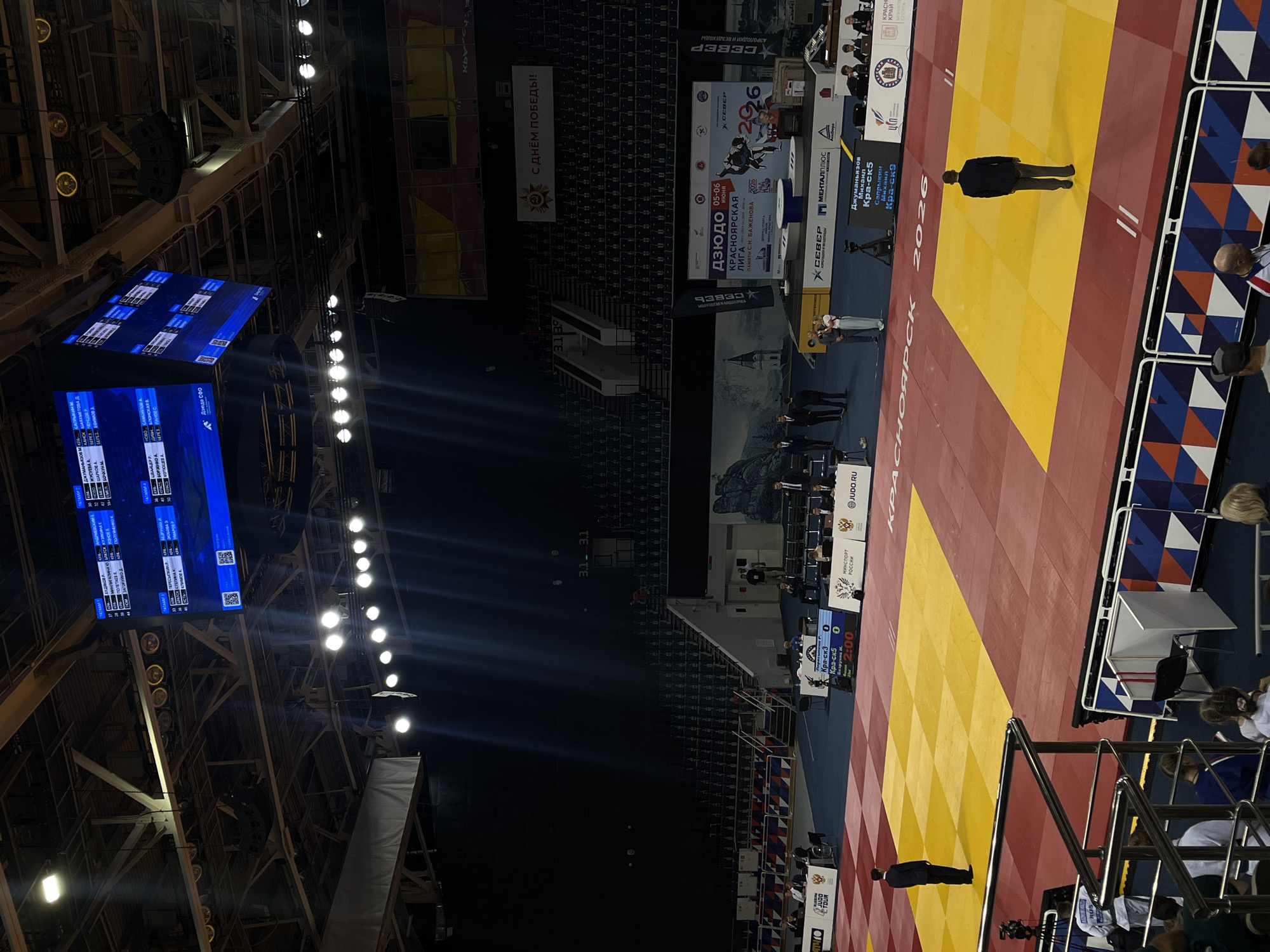 The image captures an indoor sports event, likely a judo competition, held in Krasnoyarsk, Russia. The setting is a large, dimly lit arena with high ceilings and visible structural elements. Bright spotlights illuminate the central competition area, which features multiple judo mats (tatami).
In the foreground, a red and yellow judo mat, designated "ТАТАМИ 2," is prominent, with "КРАСНОЯРСК 2026" visible on its surface. A person in dark attire, possibly a referee or official, stands centrally on this mat. To the right, another mat area with geometric patterns in blue, red, and white is partially visible, along with spectators and other officials near the mat.
Above the competition area, large blue electronic scoreboards display information, including participant names like ПОЛЕТАЕВ А., ЗАГОРУЙКО Д., ШАЯХМЕТОВА Д., БОРЗЕНКО А., and БЛОНСКАЯ В. Scores like "Кра-ск3 0" and "Кра-ск5 0" are visible, along with a timer showing "2:00." One scoreboard indicates "ТАТАМИ 2" and another "ТАТАМИ 4," suggesting multiple simultaneous matches.
In the background, tiered spectator seating is visible, mostly empty or in shadow. Various banners and advertisements adorn the walls. Notable text on banners includes "С ДНЁМ ПОБЕДЫ!" (Happy Victory Day!), "ДЗЮДО КРАСНОЯРСКАЯ ЛИГА ПАМЯТИ С.Н. БАЖЕНОВА 2026" (Judo Krasnoyarsk League in Memory of S.N. Bazhenov 2026), and sponsor logos like "СЕВЕР," "МЕНТАЛПЛЮС," and "МИНСПОРТ РОССИИ." The event appears to be "Дзюдо СФО" (Judo SFO) and a banner mentions "Джуманьязов Михаил Кра-ск5" and "Сапрыкин Михаил Кра-ск9." The overall impression is a professional judo tournament taking place in the evening.