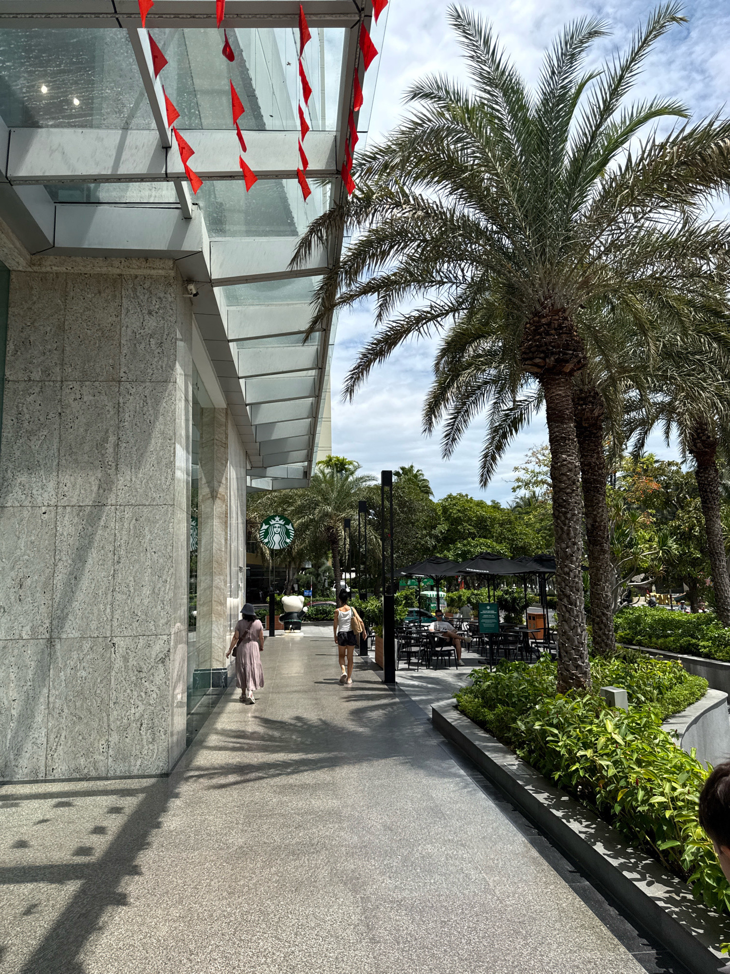 The image depicts a sunny day in Nha Trang, Vietnam, likely outside a commercial building. The foreground features a polished stone walkway with palm trees to the right, their fronds casting shadows on the ground. A modern building with a glass canopy, decorated with small red flags, stands to the left.
Two individuals are walking away from the camera on the walkway. The person in the foreground on the left is wearing a long, light-colored dress and a hat. The person to their right is wearing a white tank top and shorts. Further down the walkway, a Starbucks sign is visible, indicating a coffee shop is present. Several outdoor tables and black umbrellas for seating are set up near the Starbucks. In the background, lush green trees and foliage create a tropical atmosphere. The sky is partly cloudy.