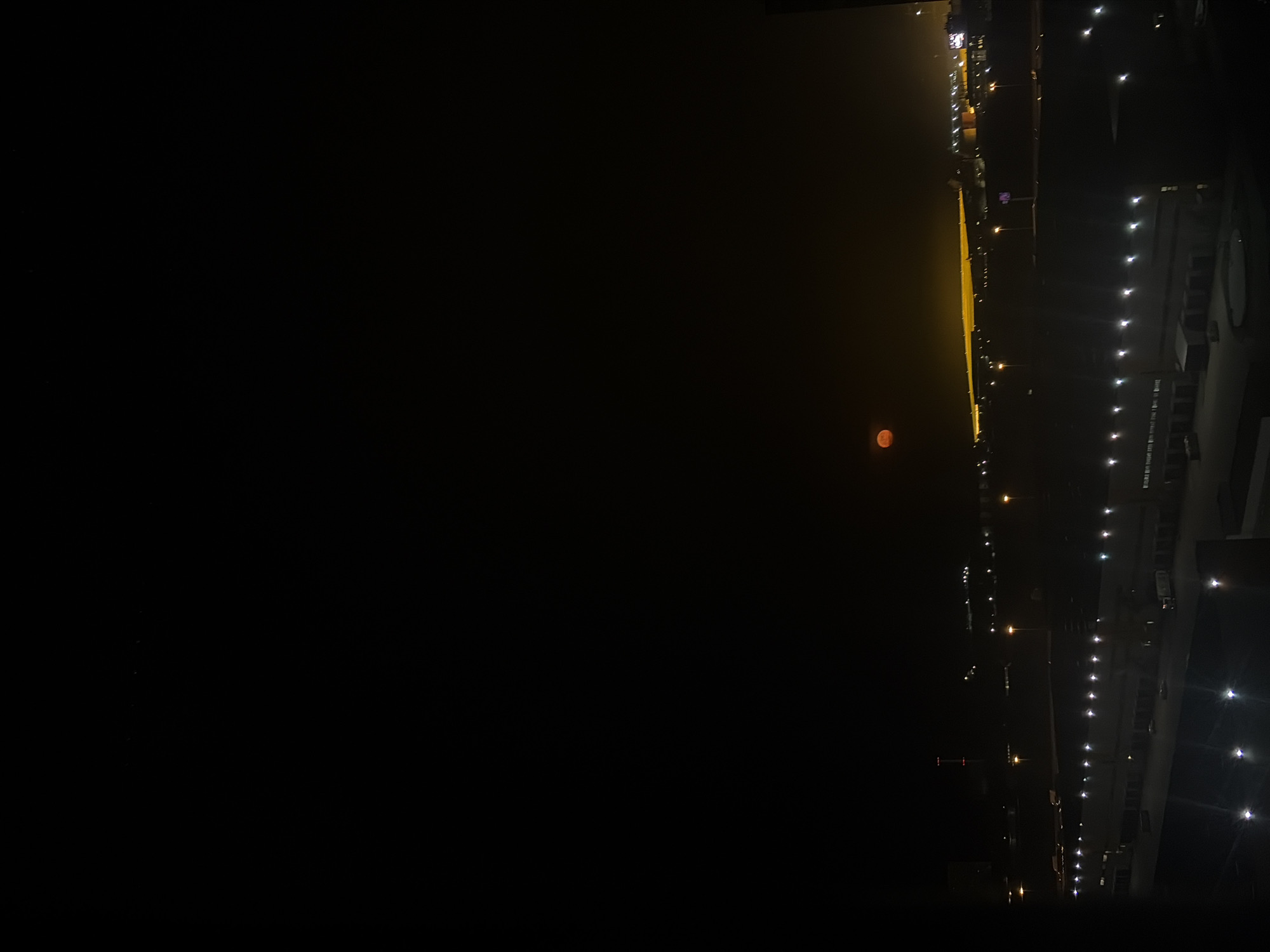 The image shows a night view of what appears to be a cityscape, potentially Saint Petersburg, Russia, based on the urban setting and possible presence of streetlights and buildings. The scene is dark with a predominantly black background. A red moon is visible in the sky. Lights from buildings and street lamps illuminate part of the cityscape, creating a stark contrast. The image is oriented sideways, with the cityscape appearing along the right side of the frame. Details are limited due to the low light conditions.