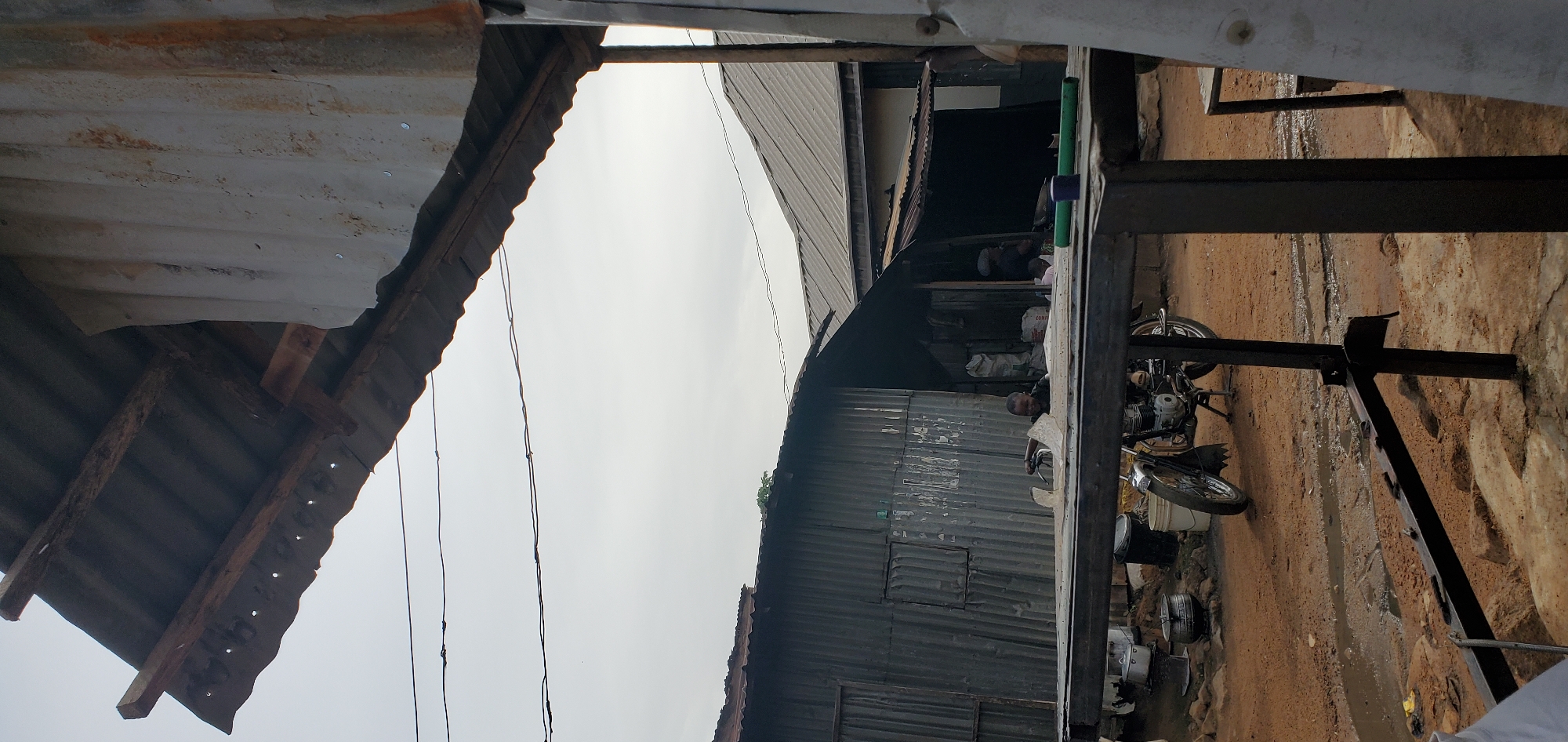 In a predominantly outdoor scene, a young boy wearing a black shirt is seated on a motorcycle. He appears to be looking towards the viewer with a slight smile. The motorcycle is positioned on a dirt ground, with puddles of water visible. Behind the boy and the motorcycle are several structures with corrugated metal roofs and walls, suggesting a rustic or informal settlement. One structure has an open doorway, revealing sacks and possibly other goods inside, and another person sitting within. Another corrugated metal structure to the left has a small window or opening. The sky is overcast. In the foreground, a dark metal structure, possibly a table or a frame, is positioned in front of the motorcycle and the boy. Overhead, electrical wires are strung across the sky. The overall impression is one of a simple, possibly rural or informal urban environment.