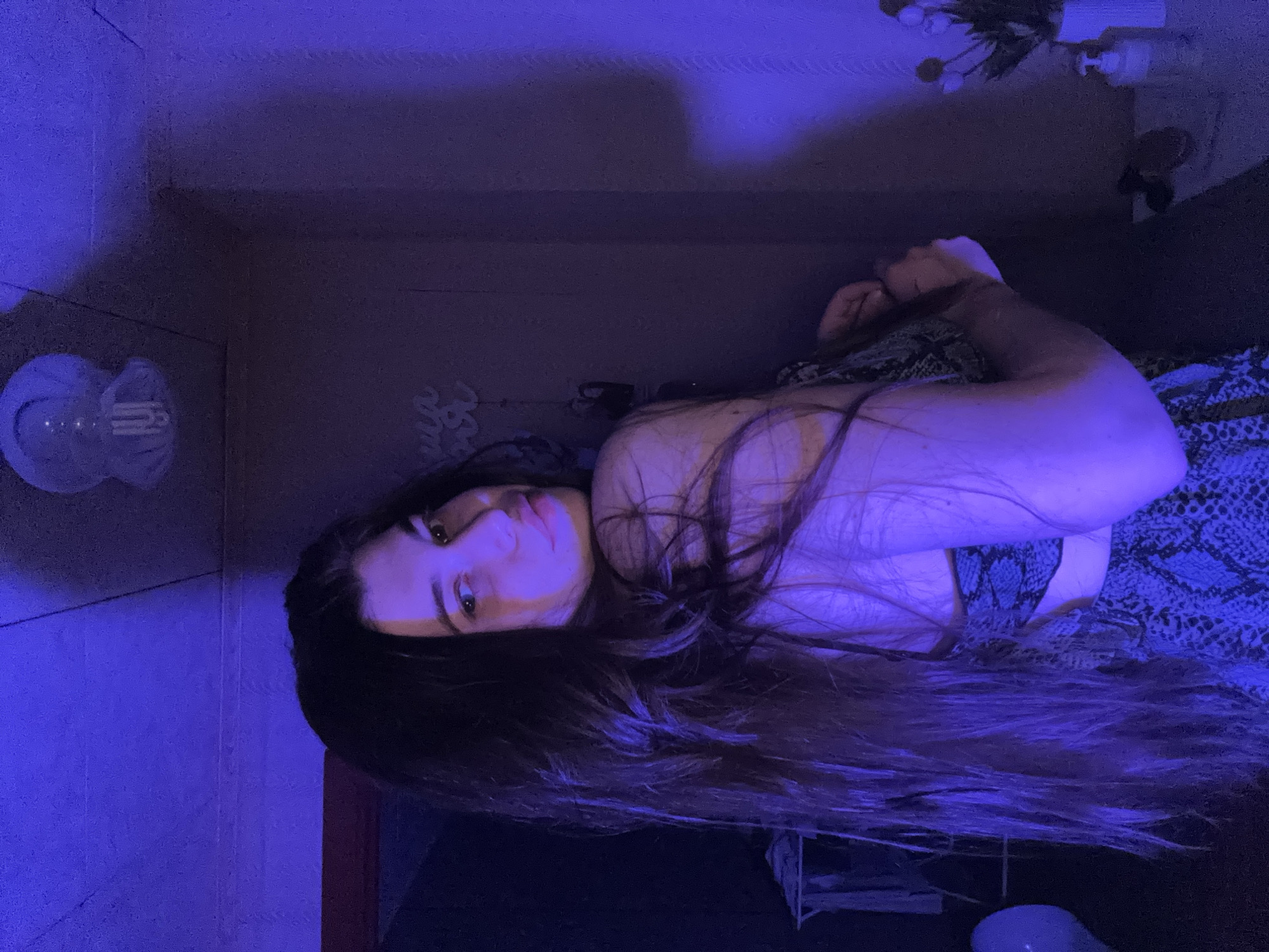 A young woman is laying down in a bathroom. The bathroom has blue lighting, and the wall tile appears to be a light brown. The woman has long, dark hair and is wearing a top with a snakeskin pattern. A toilet paper holder is visible on the wall. A vase of flowers is also visible on a ledge in the background.
