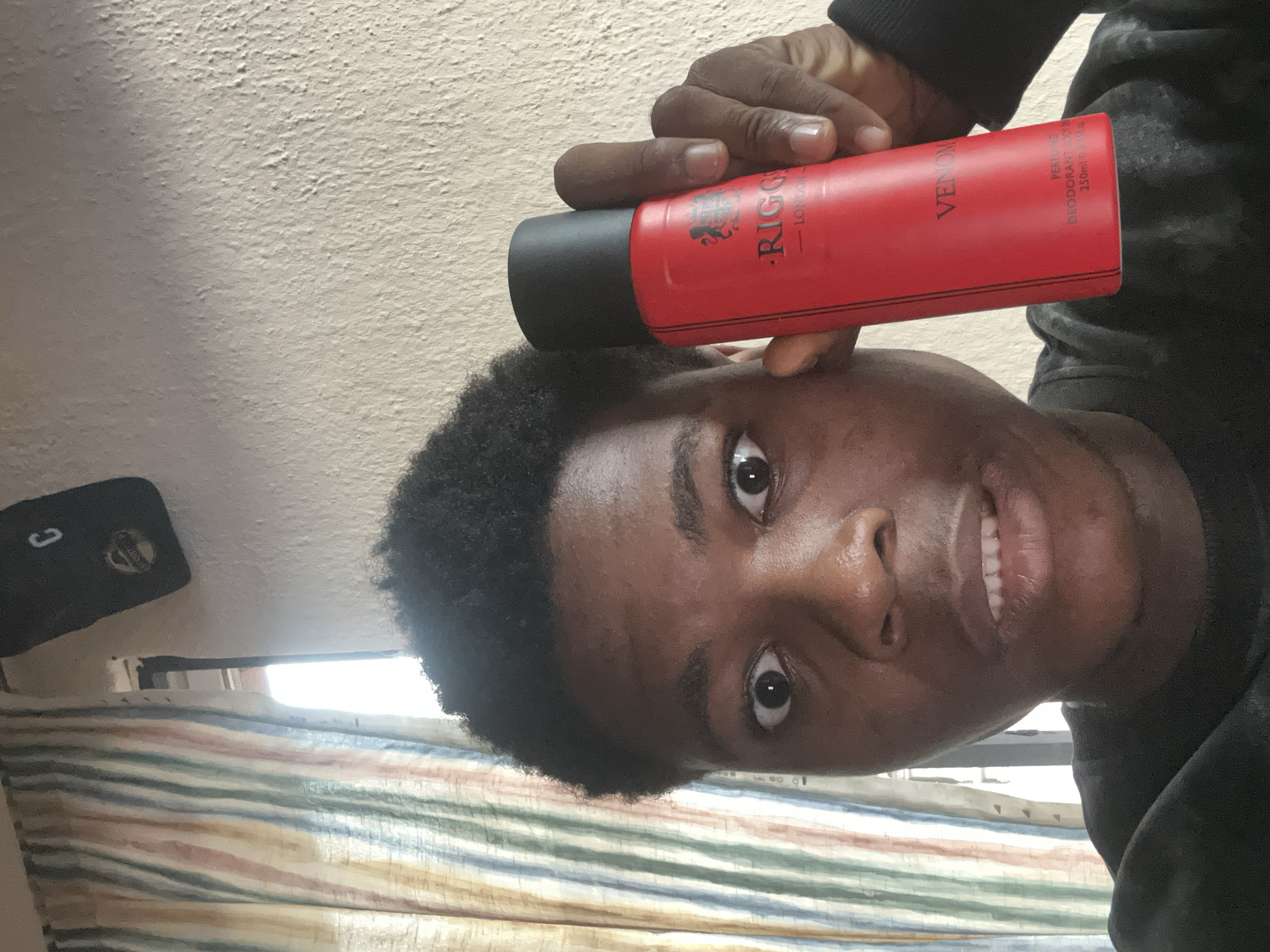 A young Black male is holding a red aerosol can of "RIGGS LONDON VENOM" deodorant. He is close to the camera, with his eyes wide open and a slight smirk on his lips. His hair is short and curly. He is wearing a black crew-neck shirt. The background shows a textured wall and a striped curtain, suggesting an indoor setting. The image has been identified as being taken in Ihiagwa, Nigeria.