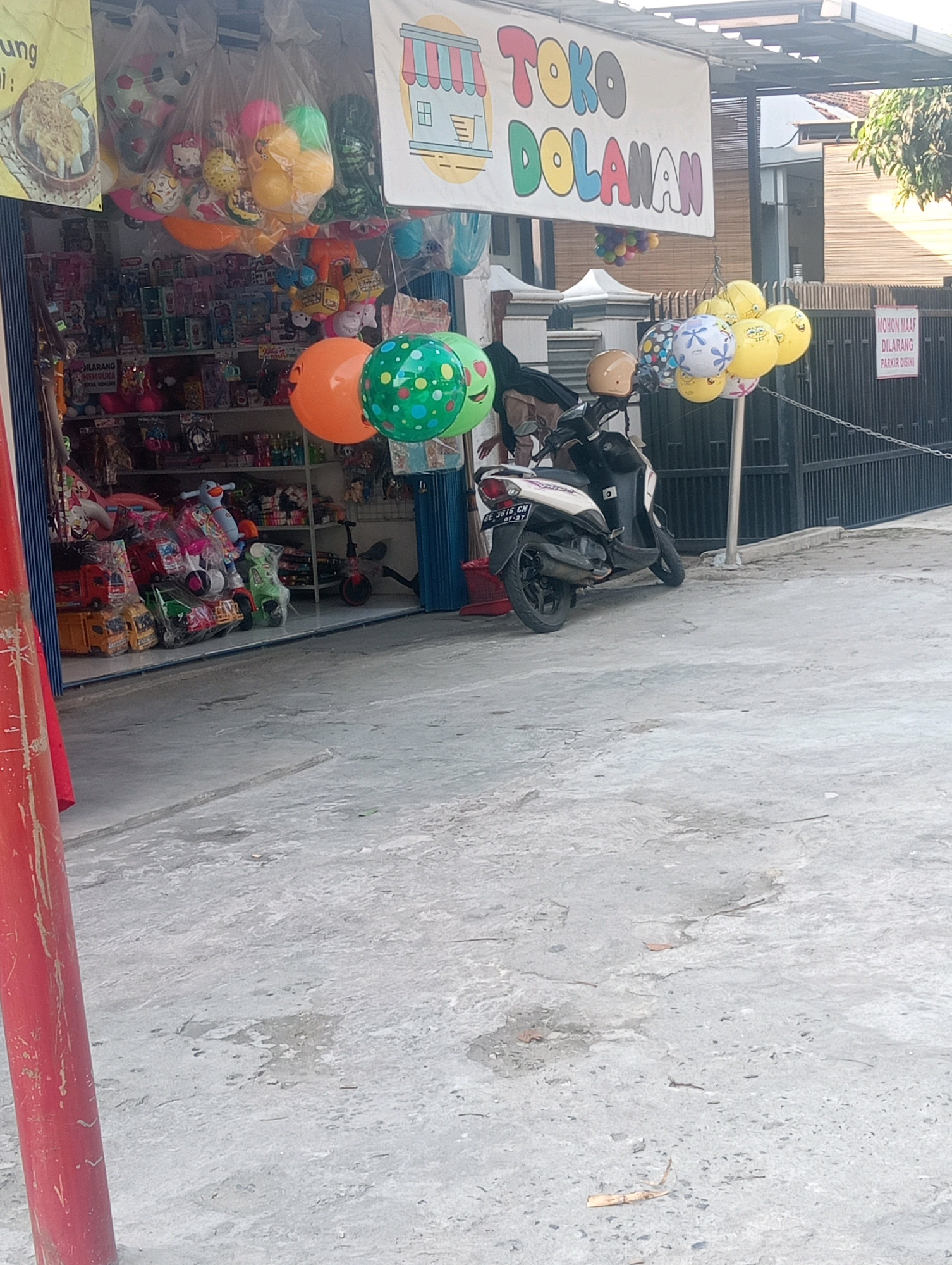 The image shows a street-level view of a toy store in Bandar Lampung, Indonesia. The storefront, labeled "Toko Dolanan," features a variety of toys displayed both inside and outside. Several balloons, including some shaped like soccer balls and cartoon characters, are hanging from the store's awning and tethered to a pole. A white scooter is parked in front of the store, with a person partially visible behind it. A sign on a metal fence behind the scooter reads "MOHON MAAF DILARANG PARKIR DISINI" (Sorry, parking here is prohibited). The ground in front of the store is paved with concrete. The overall scene depicts a typical commercial street in Indonesia, with a focus on the toy store and its offerings.