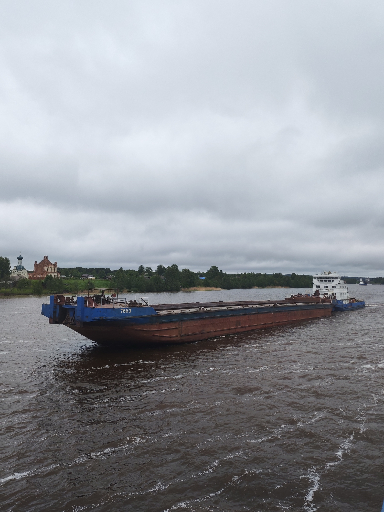 The image depicts a barge moving on a river, presumably the Volga, near Анхимово, Russia. The barge is dark brown and blue, with the number "7663" visible on its side. A tugboat is connected to the barge at its rear. The water is dark and choppy. On the riverbank to the left, a church complex with distinctive onion domes and a brick structure is visible amidst green trees and grass. The sky is overcast, creating a subdued lighting condition.