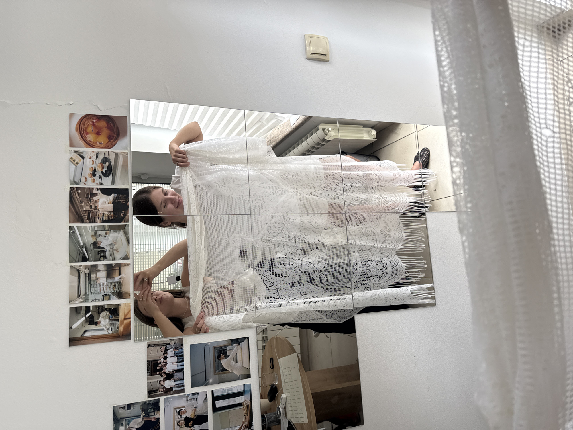 The image displays a wall decorated with various photographs and a large mirrored surface, possibly in a home or studio. In the mirror, a young woman with dark hair is visible. She is wearing a white top and appears to be holding up a piece of white lace fabric, which obscures much of her body and the reflection of the room behind her. Her facial expression suggests a thoughtful or perhaps amused demeanor.
On the wall to the left of the mirror, a stack of small, vertically oriented photographs are arranged. These photos appear to show interior scenes, possibly of kitchens or workspaces, with glimpses of appliances and people working.
Below the mirror, another cluster of photographs is present. One of these photos shows a group of people, many wearing white shirts, standing together. Other photos in this section are also small and depict interior scenes.
Further down on the wall, to the left, are more stacked photographs, similar in style to those above the mirror, featuring interior views.
The mirrored surface itself reflects a radiator and what appears to be a white curtain or blinds on the right side of the image. A light switch is visible on the white wall above the mirror. The overall impression is a personal and decorative arrangement, possibly capturing memories or inspirations. The lighting suggests it is daytime, and the setting appears to be indoors, in a residential or creative space.