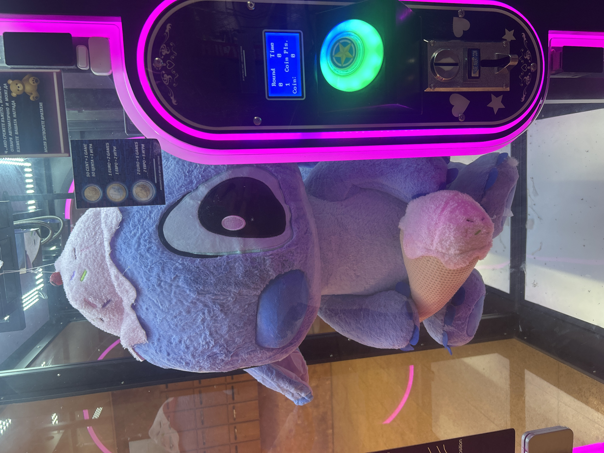 The image captures a large, light purple Stitch plush toy, wearing a pink cupcake-shaped hat with a red cherry on top and holding a pink ice cream cone, positioned inside a claw machine. The machine's right side, illuminated by vertical pink neon strips, features a control panel. This panel includes a blue-lit digital display screen showing "Round 0," "Time 0," "1 Coin Pls.," and "Coin: 0." Below the screen is a prominent, circular green-lit button with a yellow star emblem at its center. Further down is a metallic coin slot with the embossed text "PLEASE INSERT COIN."
Adhering to the machine's glass, a white sticker displays pricing in both English and Bulgarian: "50 CENT = 1 GAME / 50 ЦЕНТА = 1 ИГРА," "1 EURO = 2 GAMES / 1 ЕВРО = 2 ИГРИ," and "2 EURO = 5 GAMES / 2 ЕВРО = 5 ИГРИ." A smaller line below reads "МАШИНАТА НЕ ВРЪЩА РЕСТО!!!" (THE MACHINE DOES NOT GIVE CHANGE!!!). Above this, another sign features a small brown teddy bear and Bulgarian text which translates to instructions for retrieving a prize and a request to close the door.
The interior of the machine has visible internal lighting, and the background beyond the machine appears to be an indoor public space, possibly a mall or arcade, indicated by blurred reflections on the glass and indistinct architectural elements. The currency and language combination (Euro, Bulgarian) indicates the location is in Sofia, Bulgaria. No individuals are visible in the frame.