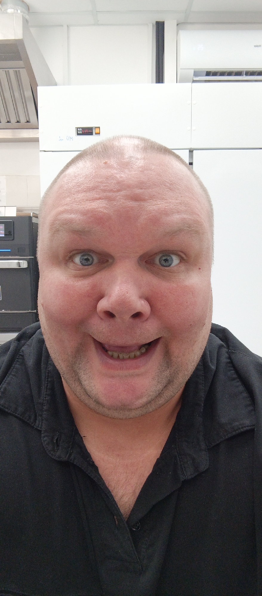 A close-up, head-and-shoulders selfie depicts a Caucasian male with a bald head and blue eyes, who is smiling widely. He is wearing a black collared shirt. The background appears to be an indoor commercial or industrial setting with white walls and some visible equipment, including a silver ventilation hood on the left and an air conditioning unit on the right. The specific location within Bugry, Russia, cannot be determined from the provided image.