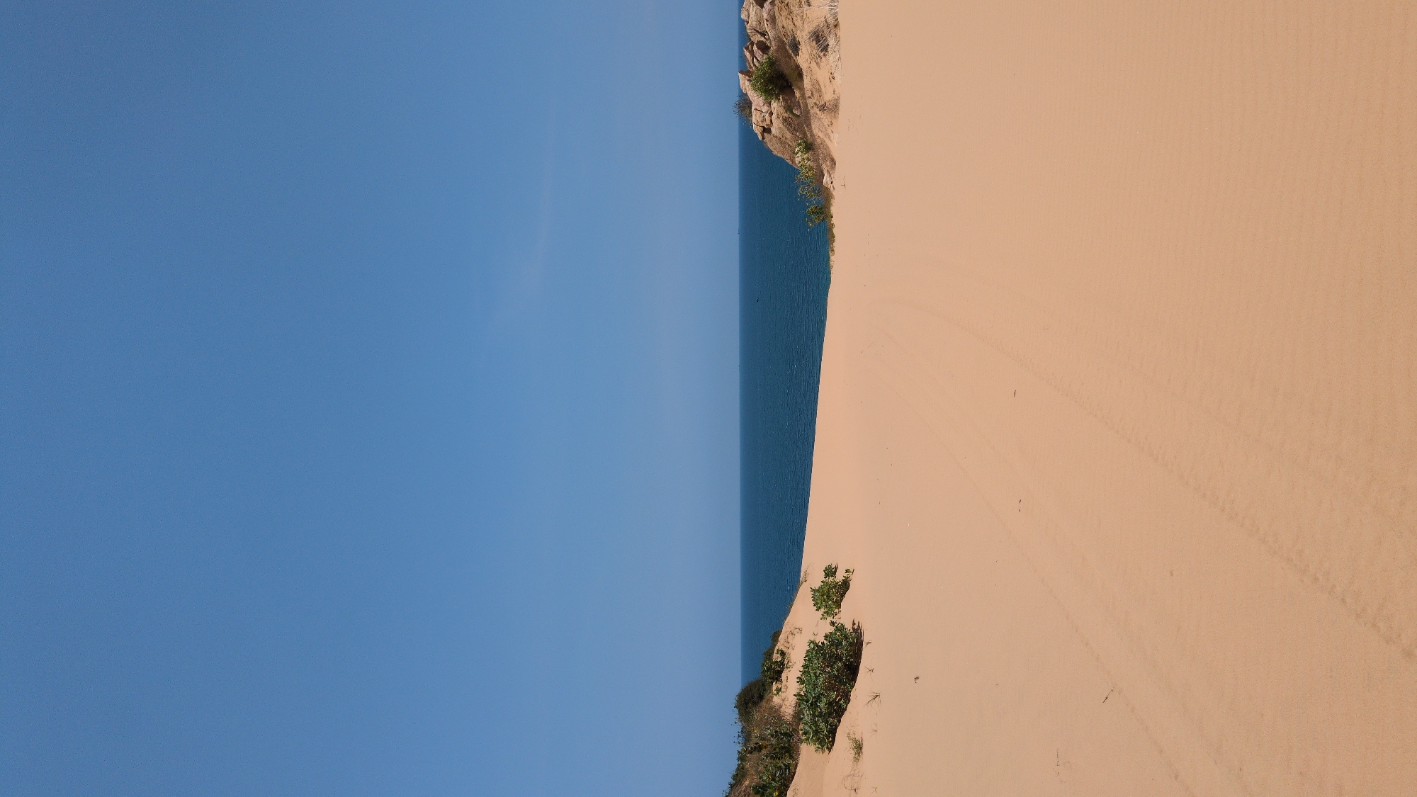 The image depicts a coastal scene in Phuoc Dinh, Vietnam. The left side shows a clear, blue sky transitioning into a band of dark blue water. To the right, a tan-colored sand dune slopes down to meet the water. Sparse vegetation grows on the dune near the bottom of the image, while the top shows a rocky outcrop with more vegetation clinging to it. There are tire tracks visible on the sand dune.