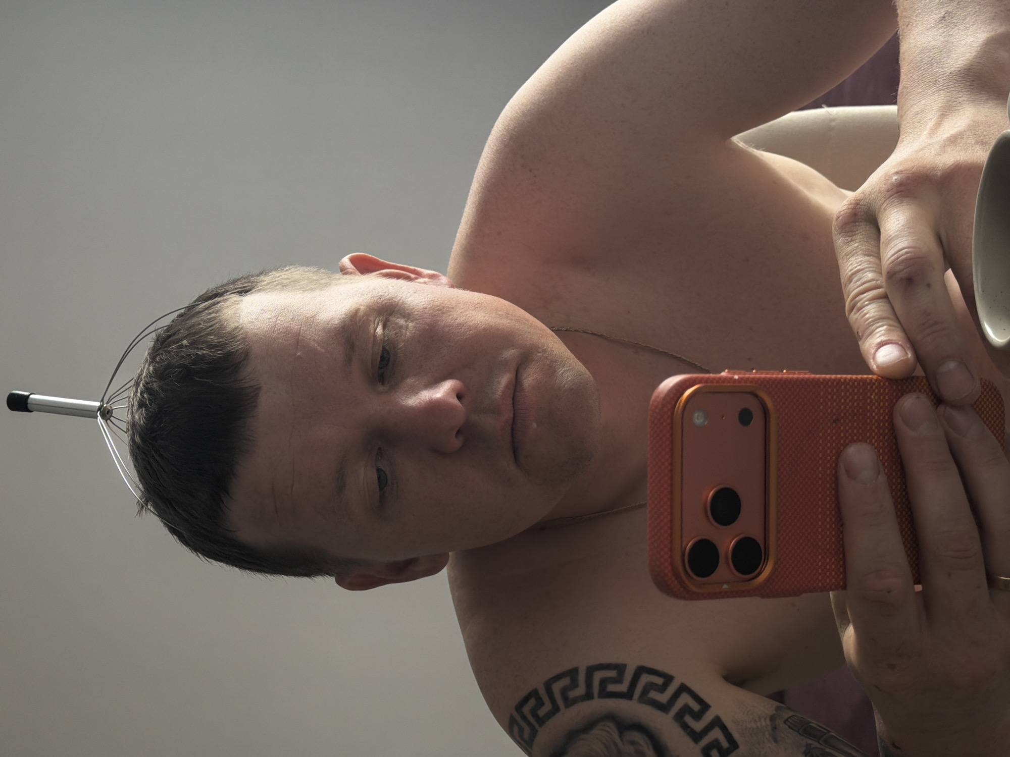 A man is looking at his phone while using a head massager. He is shirtless and has a tattoo on his left shoulder. The man is holding an orange-colored phone with three black camera lenses. He is sitting at a table, and a mug is visible in the foreground. The background is a plain grey wall. The image depicts a casual, indoor scene. The location in Chaikovsky, Russia cannot be determined from the image alone.
