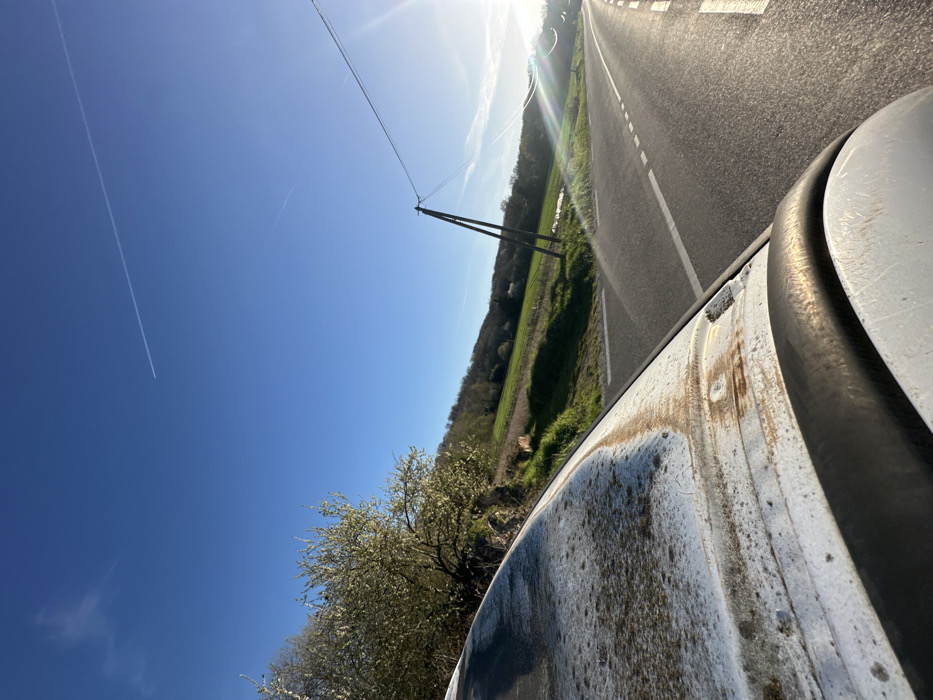 The image displays a daytime, outdoor scene in Saulx-les-Chartreux, France, captured from a low perspective over the roof of a white vehicle. The immediate foreground features the grimy, weathered white roof of the vehicle, showing areas of moss and distinct brown-orange rust streaks within its channels. A black rubber seal borders the visible edge of the roof.
In the midground, a paved asphalt road curves gently to the right and into the distance. The road is marked with a solid white line on its right shoulder and a broken white line in its center, indicating a two-way thoroughfare. To the left of the road, a green grassy verge transitions into a sloping hillside. This hillside is densely covered with trees, several of which exhibit white blossoms, suggesting a spring or early summer season. Further up the slope, beyond the flowering trees, green agricultural fields are visible against the terrain. A wooden utility pole, equipped with multiple overhead power lines, stands on the right side of the road.
The background consists of a clear, brilliant blue sky. The sun is a strong source of light in the upper right quadrant, producing noticeable lens flare across the scene. Two aircraft contrails are present in the sky: a prominent, long white contrail stretches horizontally across the upper frame, and a fainter, shorter contrail is located below and to its left. No individuals or specific human actions are depicted. The scene is static, illuminated by direct natural sunlight.