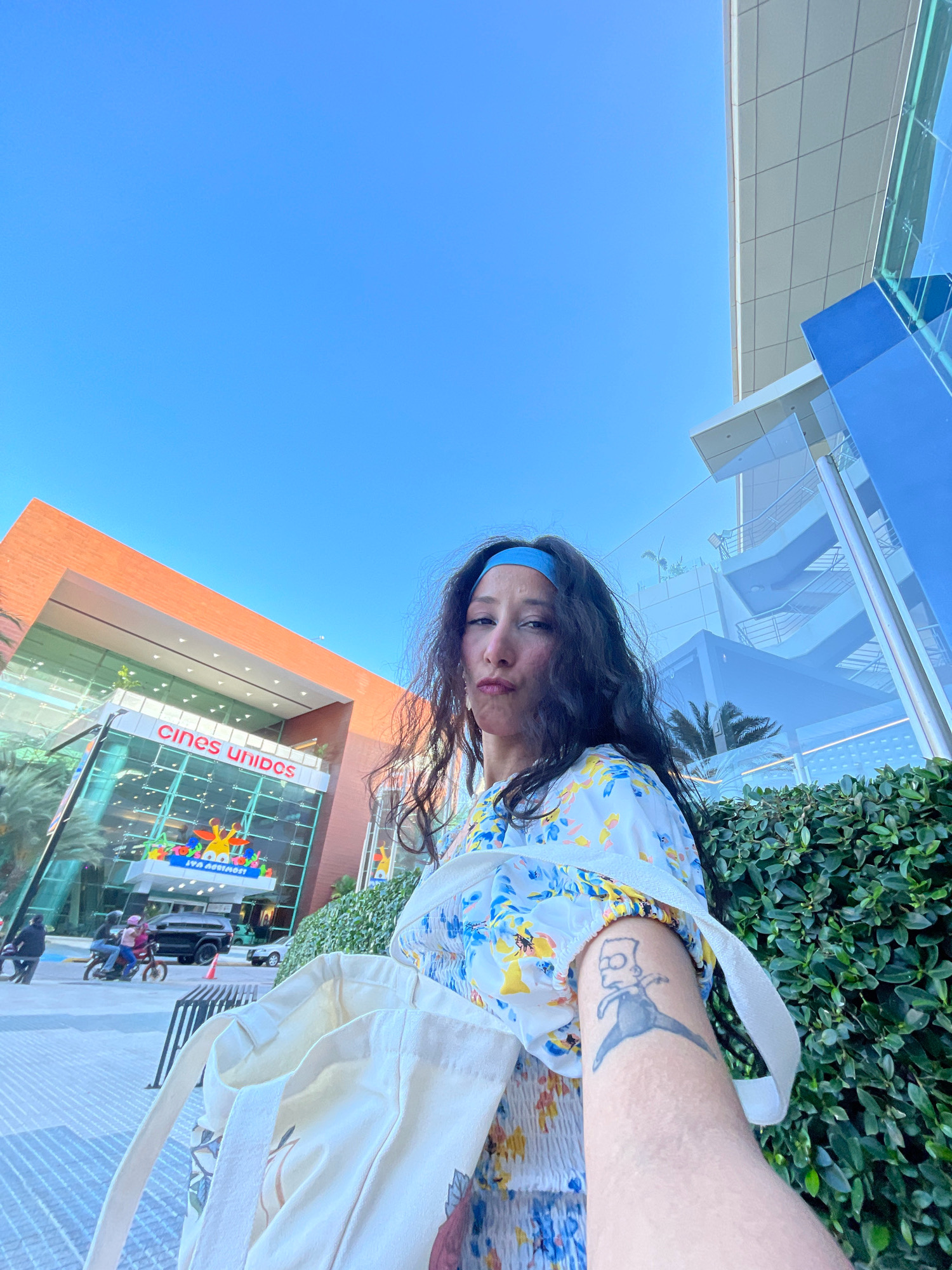 A woman with long dark hair, wearing a blue headband and a white floral dress, poses for a selfie outdoors. She is holding a canvas tote bag and has a tattoo of Bart Simpson on her right arm. Behind her, a modern building with a glass facade displays the "Cines Unidos" logo and colorful artwork. The building is flanked by a large orange structure and a manicured green hedge. The sky is clear and blue. The scene appears to be in Porlamar, Venezuela, likely near a cinema complex.