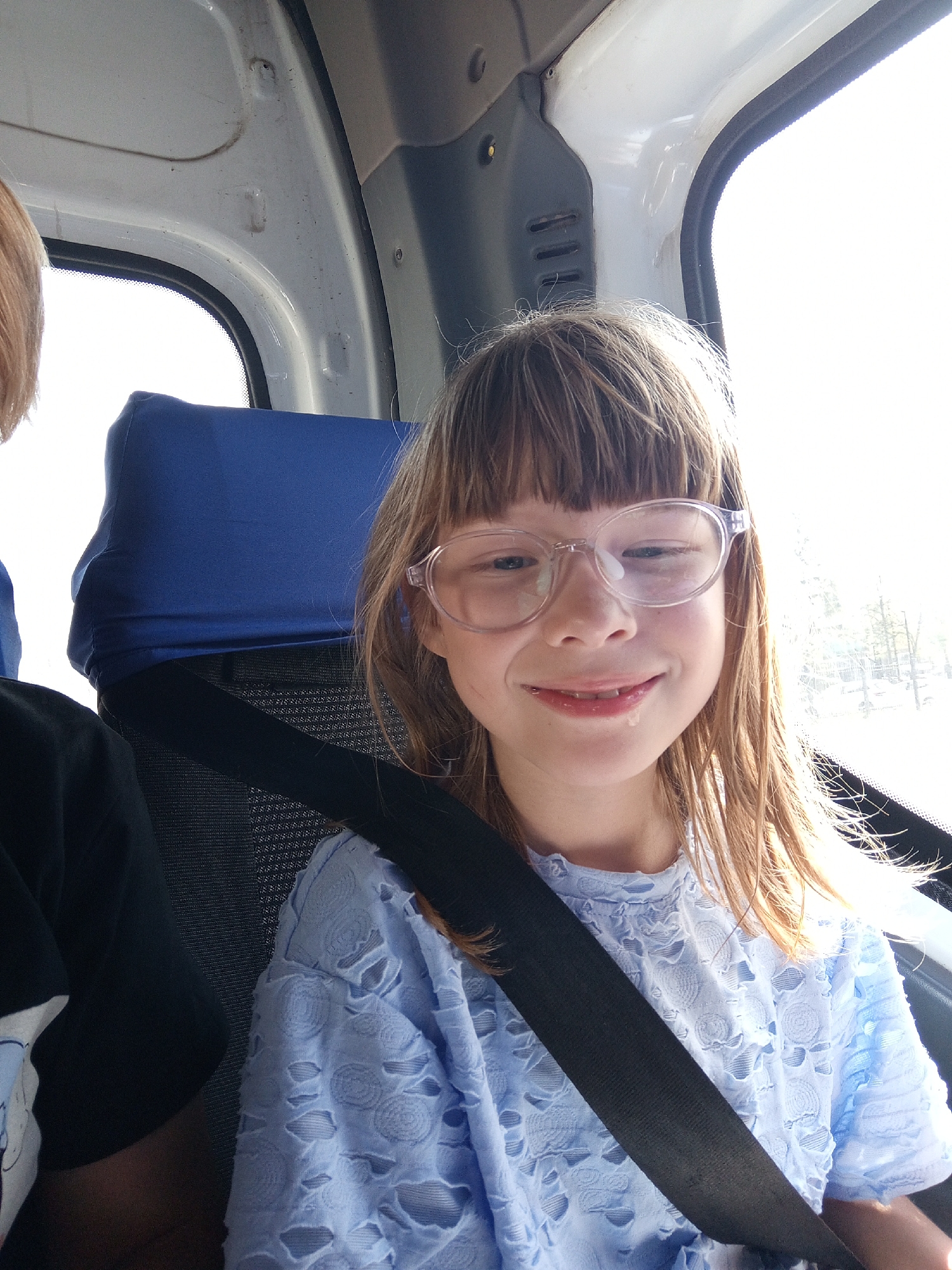 A young girl with blonde hair and bangs, wearing round, clear-rimmed glasses and a light blue, textured shirt, smiles at the camera. She appears to be in a vehicle, possibly a bus or van, as indicated by the blue headrest and seatbelt visible behind her. Her mouth is slightly open, and a small droplet of liquid is visible at the corner of her mouth. The lighting suggests it might be daytime, with bright light coming from the right side of the frame, possibly a window. The background is blurred, showing trees and a bright, overcast sky. Another person, whose head and shoulder are partially visible on the left side of the frame, is wearing a black t-shirt. The setting appears to be a casual car ride. No visible text is present in the image.