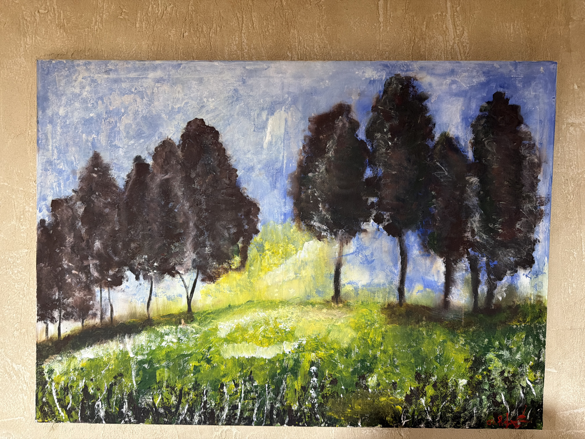 The image shows a framed oil painting depicting a landscape with trees and a grassy field. The painting is displayed on a textured wall. The style is impressionistic, with visible brushstrokes and a vibrant color palette. The foreground features a lush green field with hints of yellow and white wildflowers. Behind the field, a line of dark, silhouetted trees stands against a bright, hazy sky. The light suggests a sunny day, possibly late afternoon due to the golden hues in the middle ground. There is no discernible connection to Goristsikhe, Georgia, as the image is solely of the artwork and its immediate surroundings.