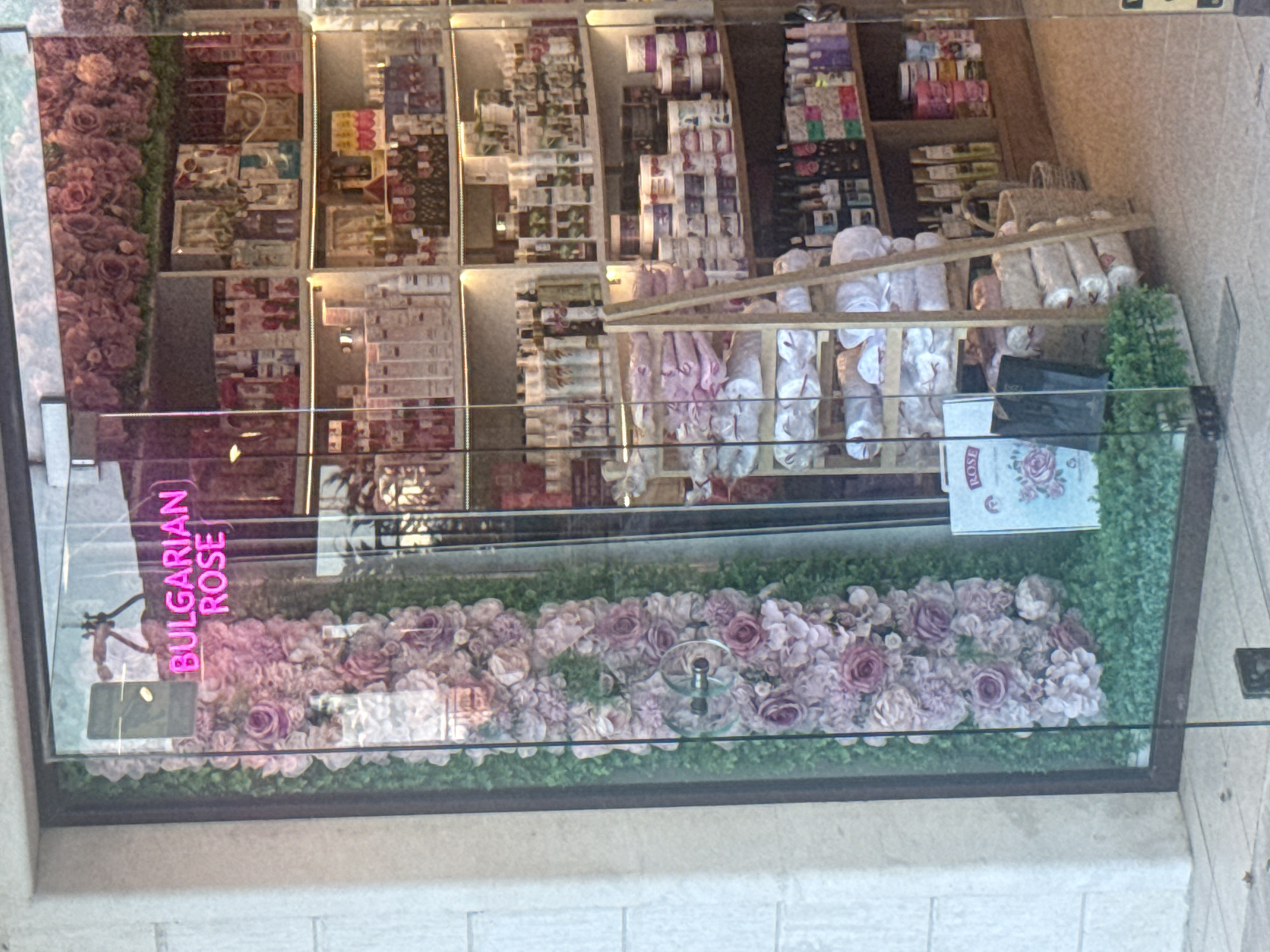 The image is a photograph of a shop window display. The window is filled with various products, likely toiletries or gifts, displayed on shelves. The backdrop features an artificial floral arrangement of pink and purple roses, interspersed with greenery. A neon sign is visible, vertically oriented, with the text "BULGARIAN ROSE" in bright pink. In the lower portion of the display, products are arranged on shelves, including what appear to be rolls of fabric or paper, packaged soaps, and boxes of cosmetics. Some of the products are wrapped in white paper or plastic. A wicker basket is also present on one of the shelves. The overall impression is of a retail display for a themed product line, emphasizing a rose scent or origin. The lighting suggests it is daytime, though the interior lighting of the shop is also visible, creating reflections on the glass.
