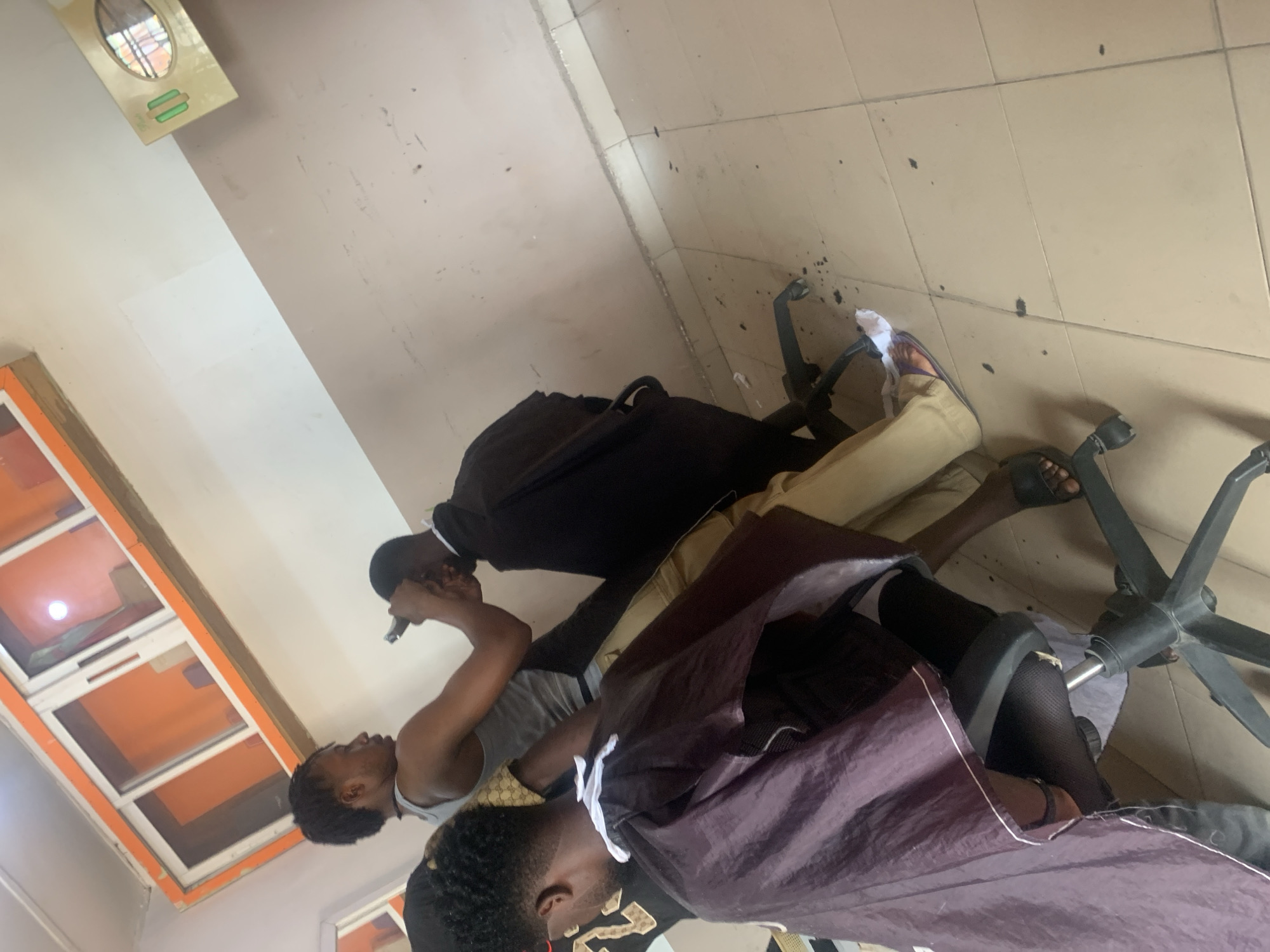 This image depicts a barber shop located in Jalingo, Nigeria. In the foreground, a young man is seated in a black barber chair, draped with a dark purple cape. He is wearing black stockings. Another young man, the barber, stands behind him, operating hair clippers to cut the seated man's hair. The barber is wearing a grey tank top and tan pants. A third individual, partially visible, is seated to the right, also draped in a cape. The environment includes a tiled floor with scattered debris, and a wall with cabinetry above. The overall scene suggests a typical barbershop service in progress.