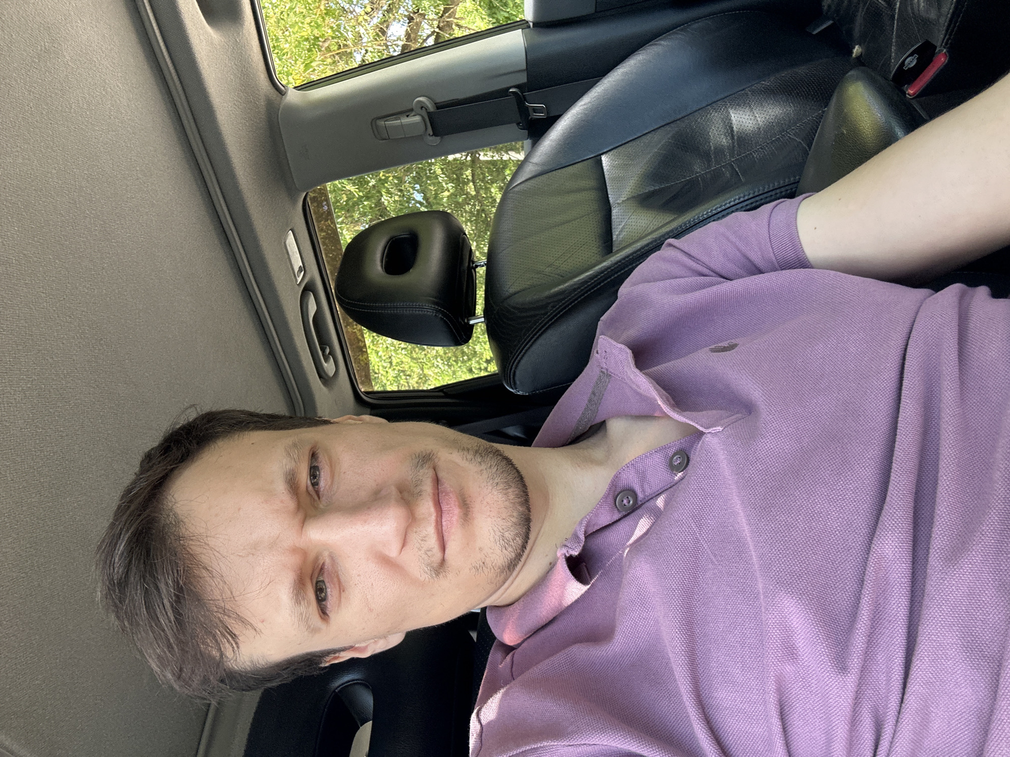 A man with short, dark hair and stubble is in the driver's seat of a car, taking a selfie. He is wearing a purple polo shirt with two buttons. The interior of the car is visible, including a black leather seat and headrest, a seatbelt, and the car's ceiling. Sunlight is streaming in through the windows, illuminating the scene and casting shadows. The background shows greenery, suggesting an outdoor location, possibly in a rural or suburban area. The specific location in Makiv, Ukraine, cannot be determined from the image alone.
