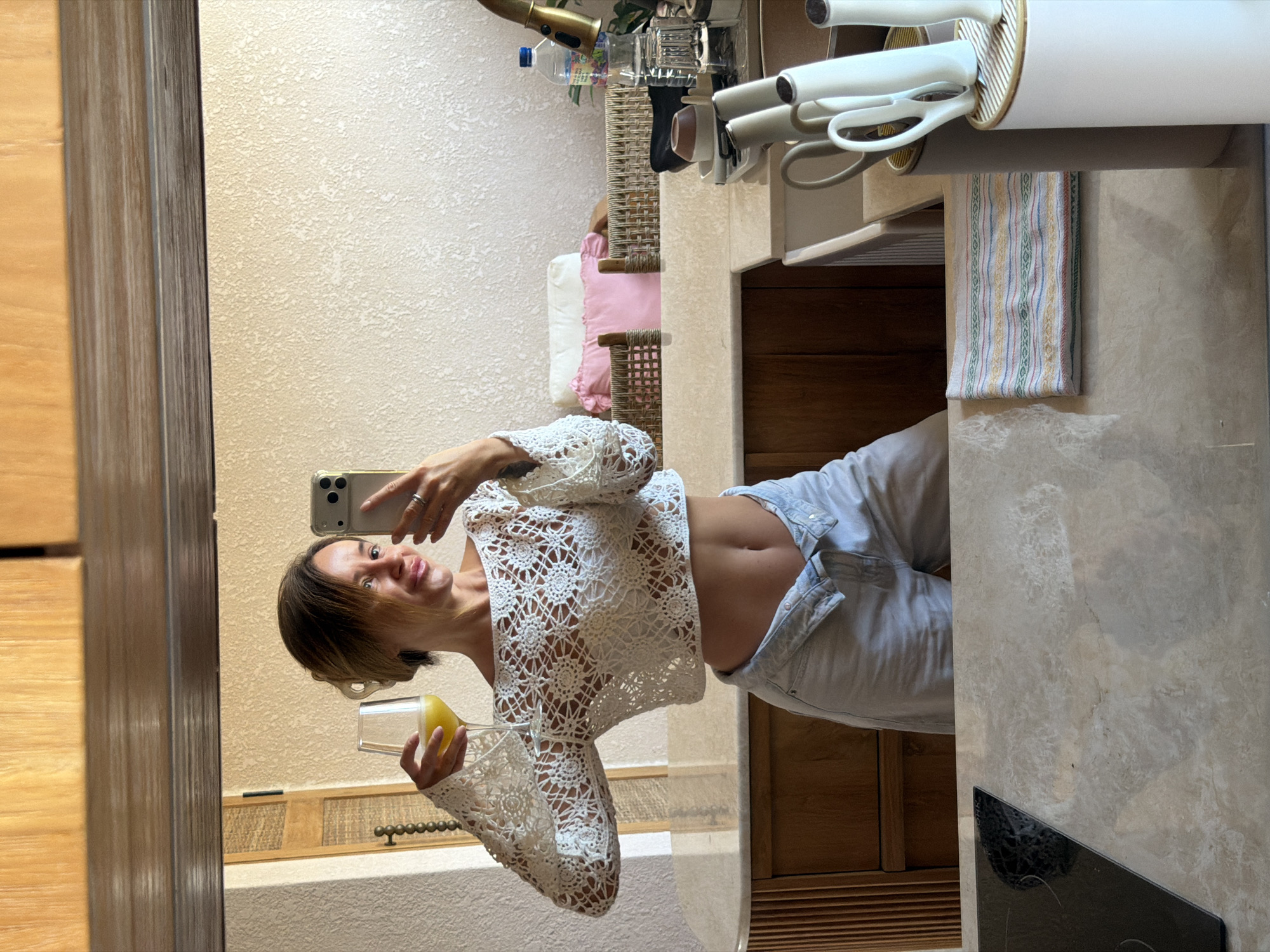 A woman is taking a selfie in a mirror in a kitchen. She is wearing a white crocheted crop top and light blue denim shorts. She is holding a glass of orange juice in her left hand and her phone in her right hand. The kitchen has marble countertops and wooden cabinets. There are also some dishes and a bottle of water on the counter. The location of this image is not specified as Mengwi, Indonesia.