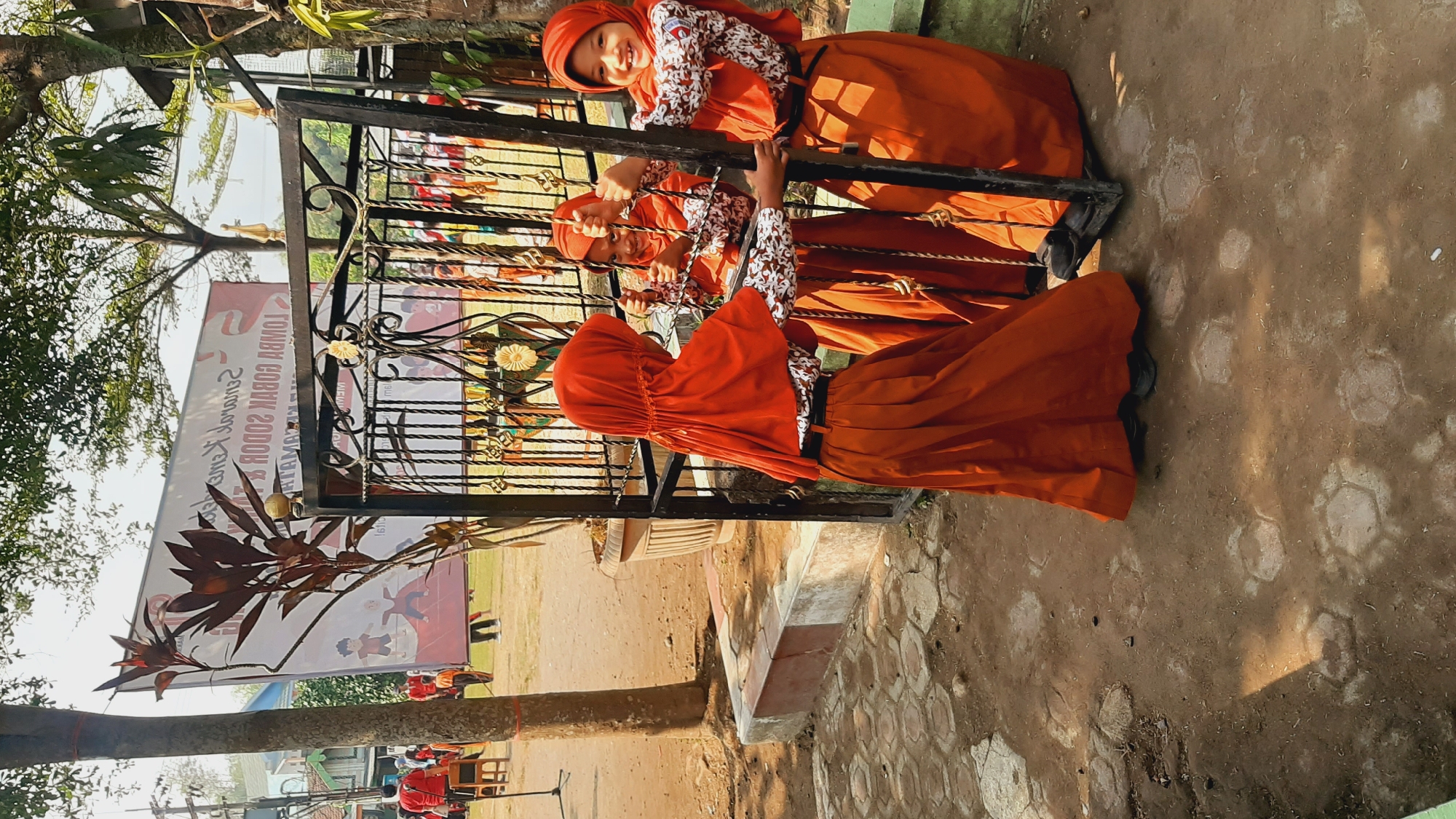 Three young girls wearing orange hijabs and matching orange skirts over patterned blouses are standing behind a black metal gate. The girl on the right is smiling widely at the camera, her hands gripping the bars of the gate. The girl in the middle is looking at the girl on the right and appears to be smiling. The girl on the left has her back to the camera. They are standing on a dirt and paver ground outside. In the background, a large banner with text and illustrations of children is visible, along with trees and other people in the distance, some wearing red. The scene appears to be in a public outdoor space, possibly a schoolyard, in Sumberjo, Indonesia.