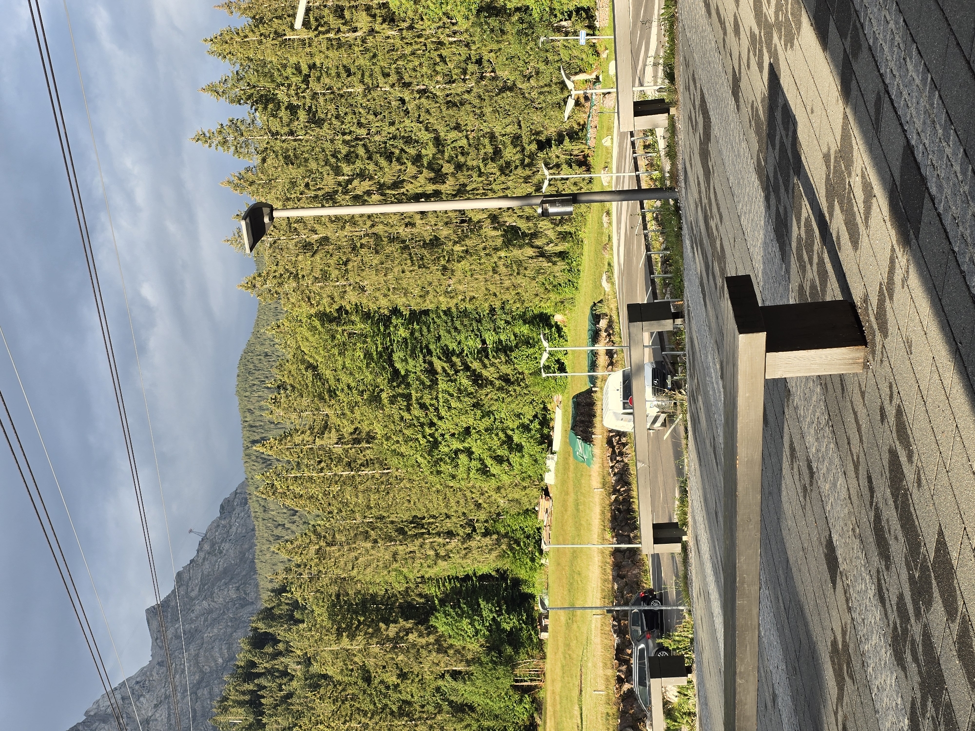 The image depicts a scene in Grainau, Germany, with a focus on the natural landscape and urban infrastructure. In the foreground, a wooden bench sits on a paved patio with wet patches, indicating recent rain or watering. Beyond the patio, a road runs horizontally, with cars parked and driving. A white van is visible on the right side of the road. A dense forest of evergreen trees forms the middle ground, extending towards a rocky mountain in the background. The sky above is cloudy, with patches of sunlight breaking through. Overhead, power lines and a cable car support structure are visible, suggesting the presence of a nearby cable car station or ski lift. The overall impression is a scenic location that blends natural beauty with functional elements of a tourist or transportation hub.