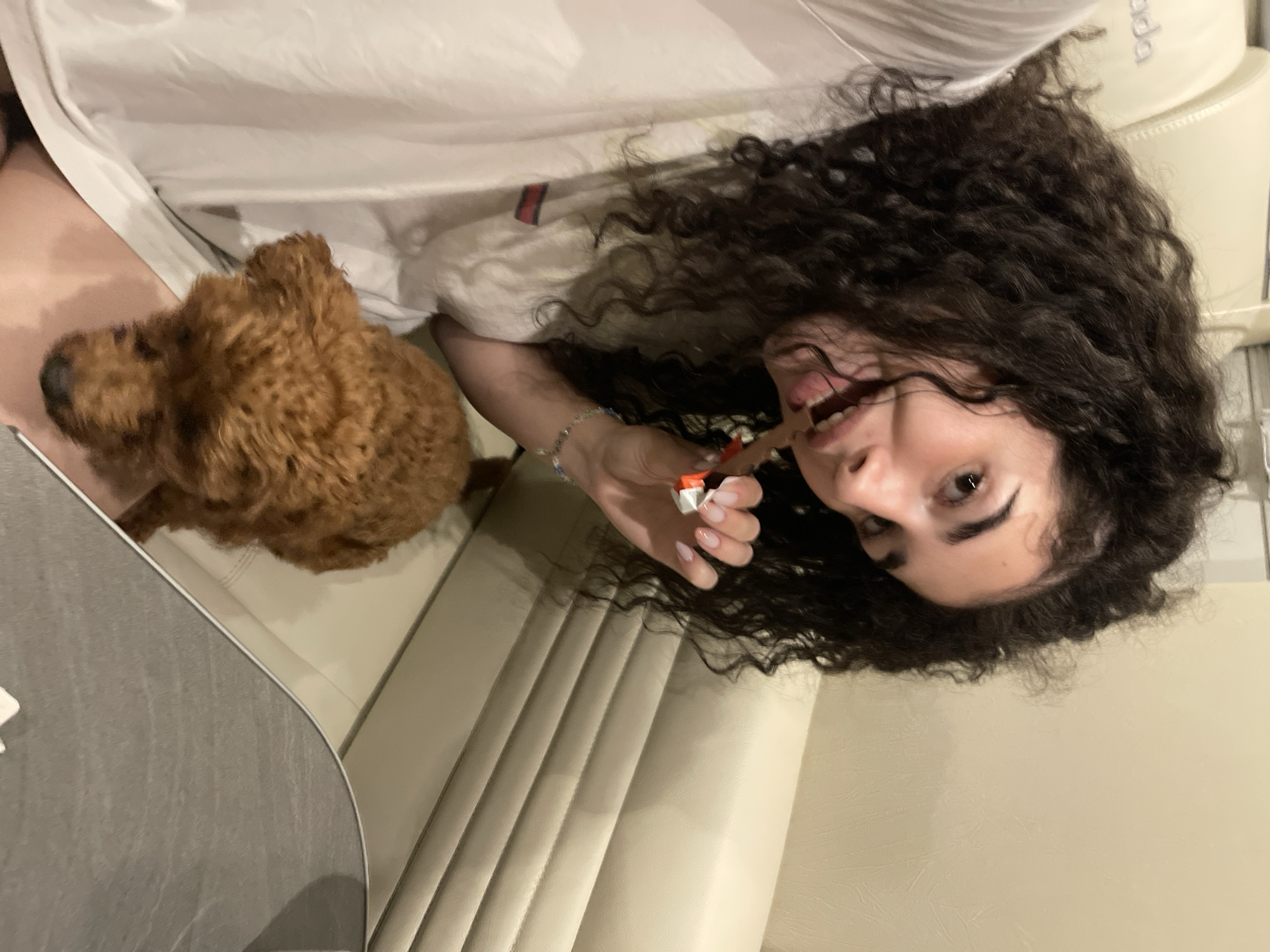 An indoor scene in Koprinka, Bulgaria, depicts a young female and a small dog. The female, with long, dark, curly hair, is positioned in the left portion of the frame, looking towards the camera with a wide smile, her mouth open and tongue visible. She wears a white short-sleeved t-shirt with a small red and blue stripe on the left shoulder/sleeve. Her left hand, adorned with a light-colored bracelet and manicured nails, holds a partially unwrapped Kinder Bueno chocolate bar near her mouth. A small, brown, curly-haired dog rests on her lap or thigh, facing downwards. The background features light-beige upholstered seating with horizontal ridges, possibly part of a sofa or car interior. A white headrest or cushion with partial dark lettering "ada" is visible in the upper left. A grey, curved surface is present in the lower right foreground.