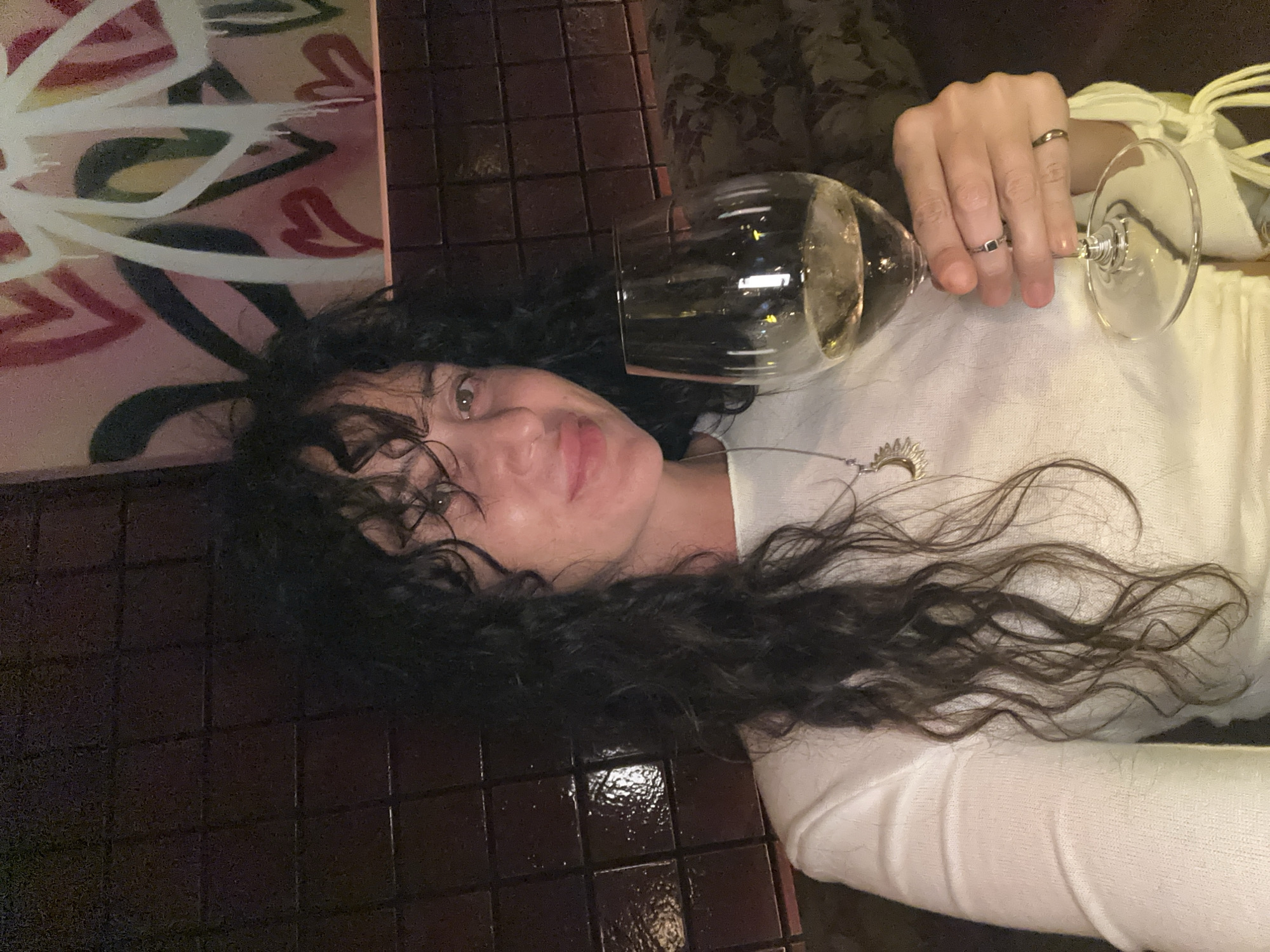 The image shows a woman holding a glass of white wine. She has long, dark, curly hair and is wearing a white top. She is also wearing a necklace with a sun pendant and two rings on her fingers. In the background, there is a wall with dark tiles and a colorful painting with graffiti art. The setting appears to be a restaurant or bar. The woman is looking at the camera and smiling slightly.