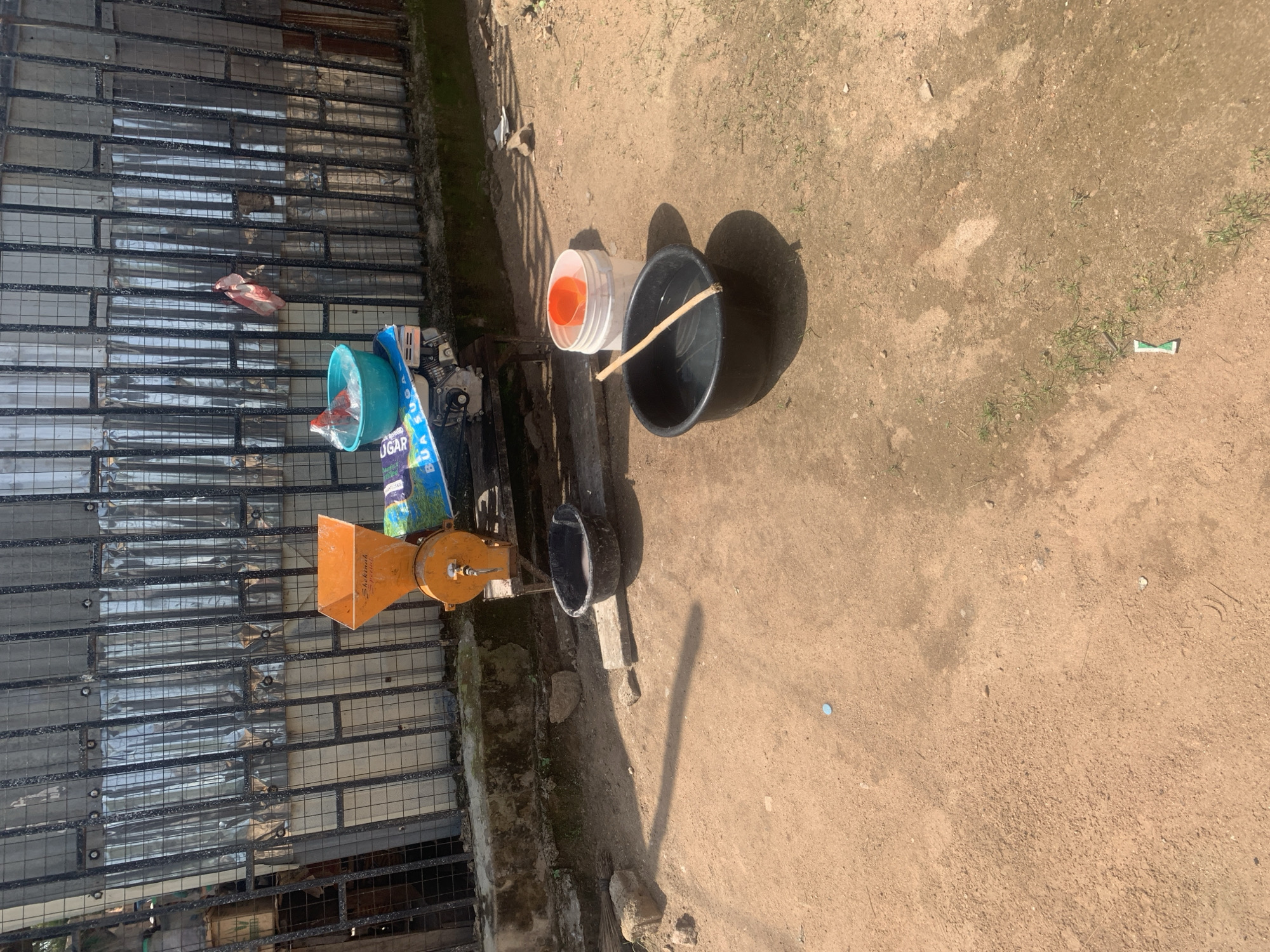 The image displays an outdoor scene, likely in a rural or semi-urban setting in Jalingo, Nigeria, during the daytime. The ground is a dusty, sandy earth with sparse patches of green grass.
Prominently featured are several objects arranged on the ground and near a wire-mesh fence. To the left, behind the fence, is a corrugated metal structure. In front of this fence, there is an orange-colored hopper with a metal spout, possibly part of a grinding machine. A blue bag with white lettering is partially visible next to the hopper.
Further to the right, a large black plastic bucket with a piece of wood lying across it is set on the ground. Next to it, a white plastic container with an orange insert is placed. A shadow from the bucket and container is cast on the ground, indicating sunlight. There are also some small pieces of debris scattered on the ground, including what appears to be a small blue cap and a white and green wrapper. The overall atmosphere suggests a practical, possibly domestic or small business setting. No people are visible in the image.