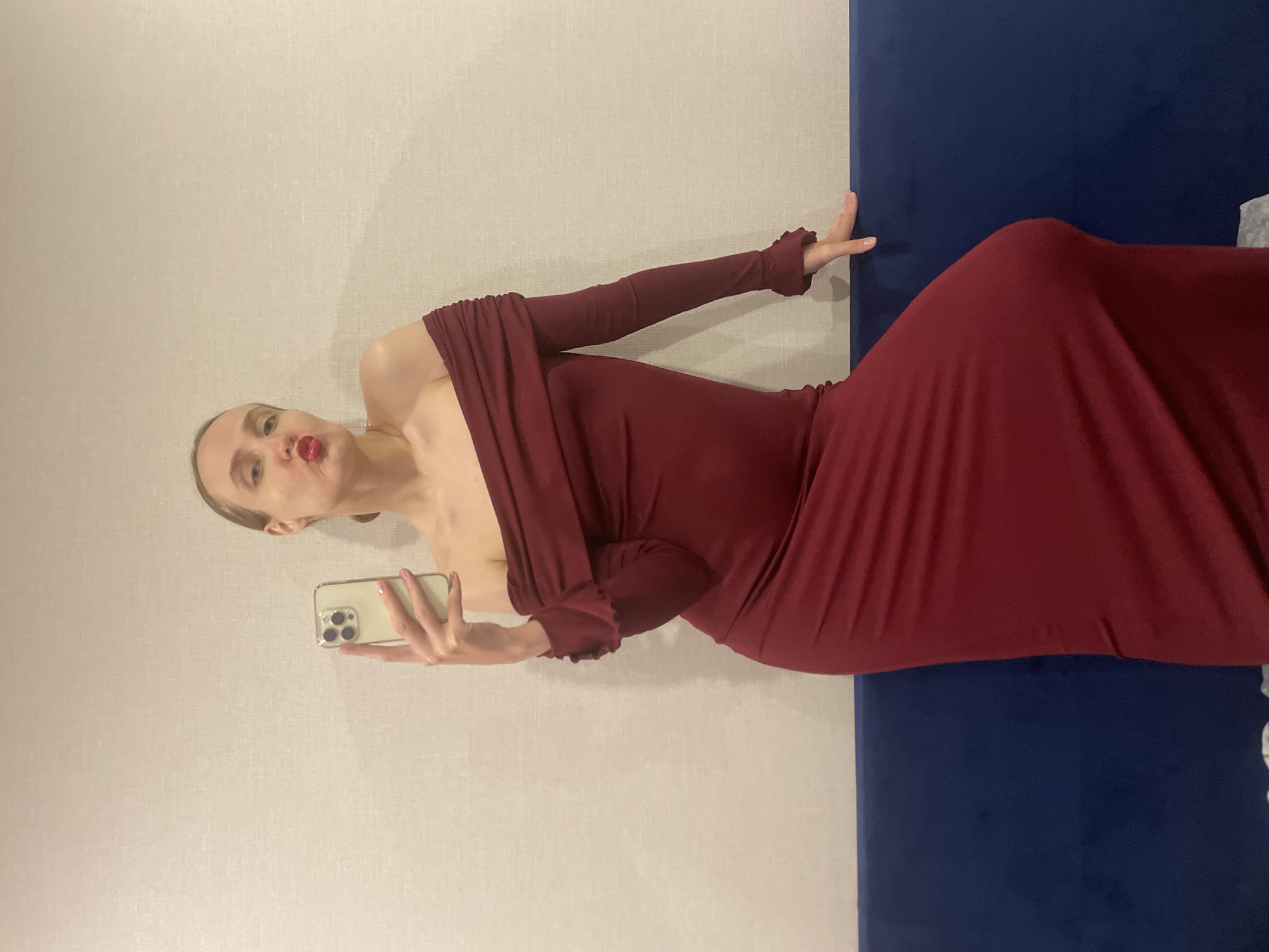 A woman is taking a selfie. She is wearing a burgundy, off-the-shoulder gown with long sleeves. She is holding a gold iPhone in her left hand. Her lips are pursed. She is positioned against a beige wall and a dark blue panel. Location: undetermined. The location "Подшивалово, Russia" cannot be confirmed from the image.