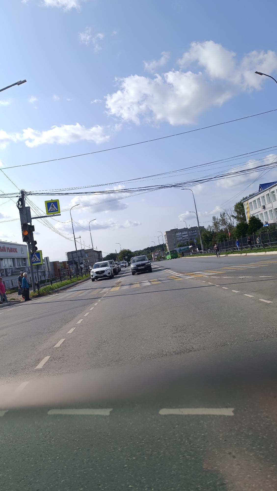 The image shows a street scene in Chaikovsky, Russia, on a cloudy day. The viewpoint is from a vehicle driving on an asphalt road with white lane markings. In the foreground, the road surface is visible with a blurred area at the bottom, likely from the vehicle's windshield.
To the left of the road, there is a pedestrian crossing with yellow warning signs featuring a pedestrian icon. A traffic light shows a yellow signal, and another sign with a pedestrian icon is below it. Adjacent to the sidewalk, a building with the inscription "ЦЕНТРАЛЬНЫЙ РЫНОК" (Central Market) is visible. Several people are standing on the sidewalk near the building.
Further down the road, there are multiple cars driving or stopped. Two pedestrian crossings are marked with yellow lines. On the right side of the road, there are more buildings, including a larger, multi-story building with a blue roof and white facade, appearing to be an administrative or commercial structure. Trees and other vegetation are visible along the roadside and in the background. Power lines are strung across the sky, supported by poles. The sky is partly cloudy with some areas of blue.