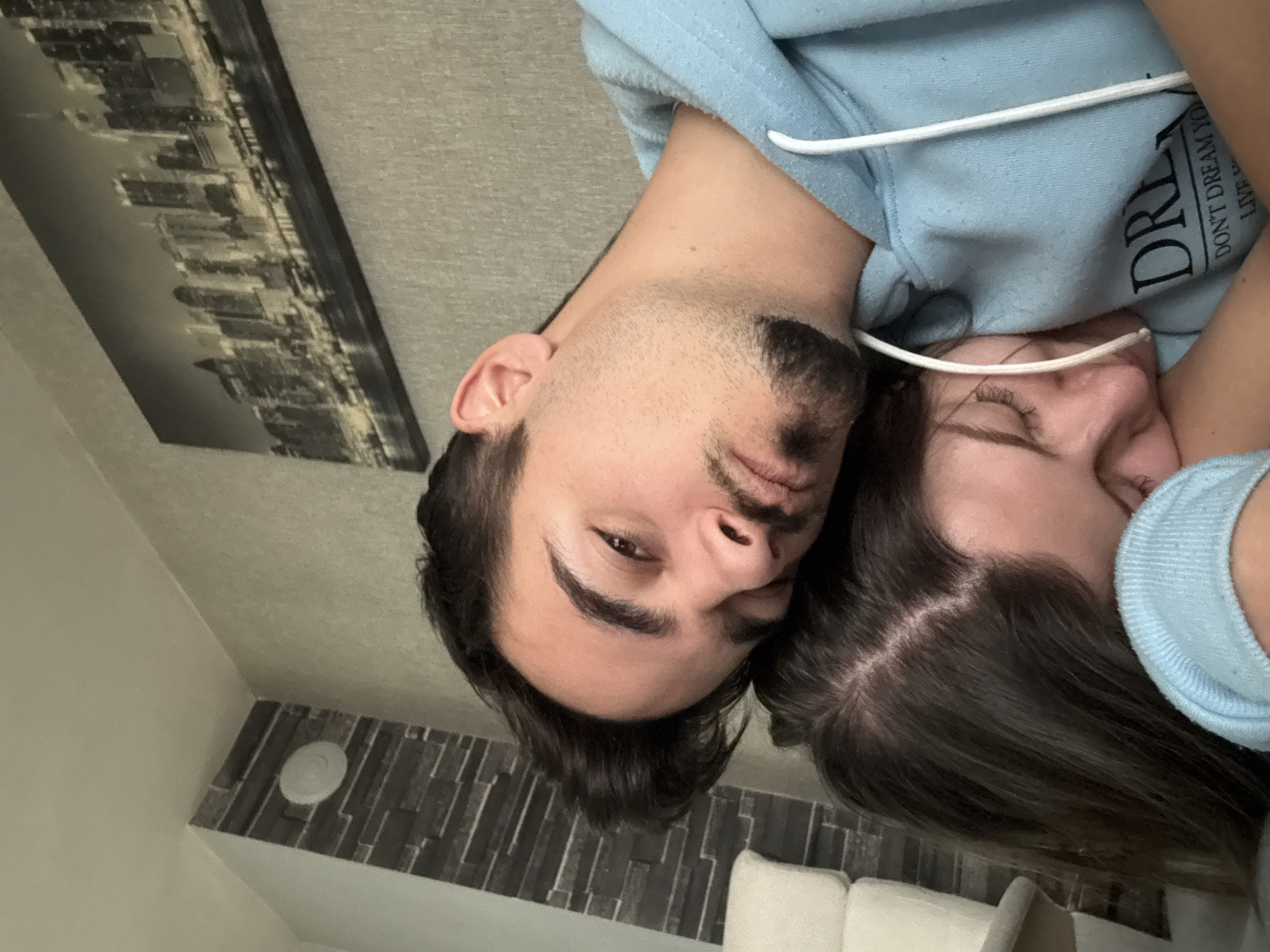 A selfie shows a man and a woman close together, possibly hugging. The man is dark-haired with a mustache and beard, and he looks directly at the camera. The woman is next to him and has her eyes closed. Both are wearing light blue hooded sweatshirts. The text on the man's hoodie reads "DREAM... DON'T DREAM YOUR LIFE, LIVE YOUR DREAM." In the background is a cityscape image in black and white and an indistinct wall area with a circular object, possibly a sensor, mounted on it. The location is most likely indoors, potentially in Sofia, Bulgaria.