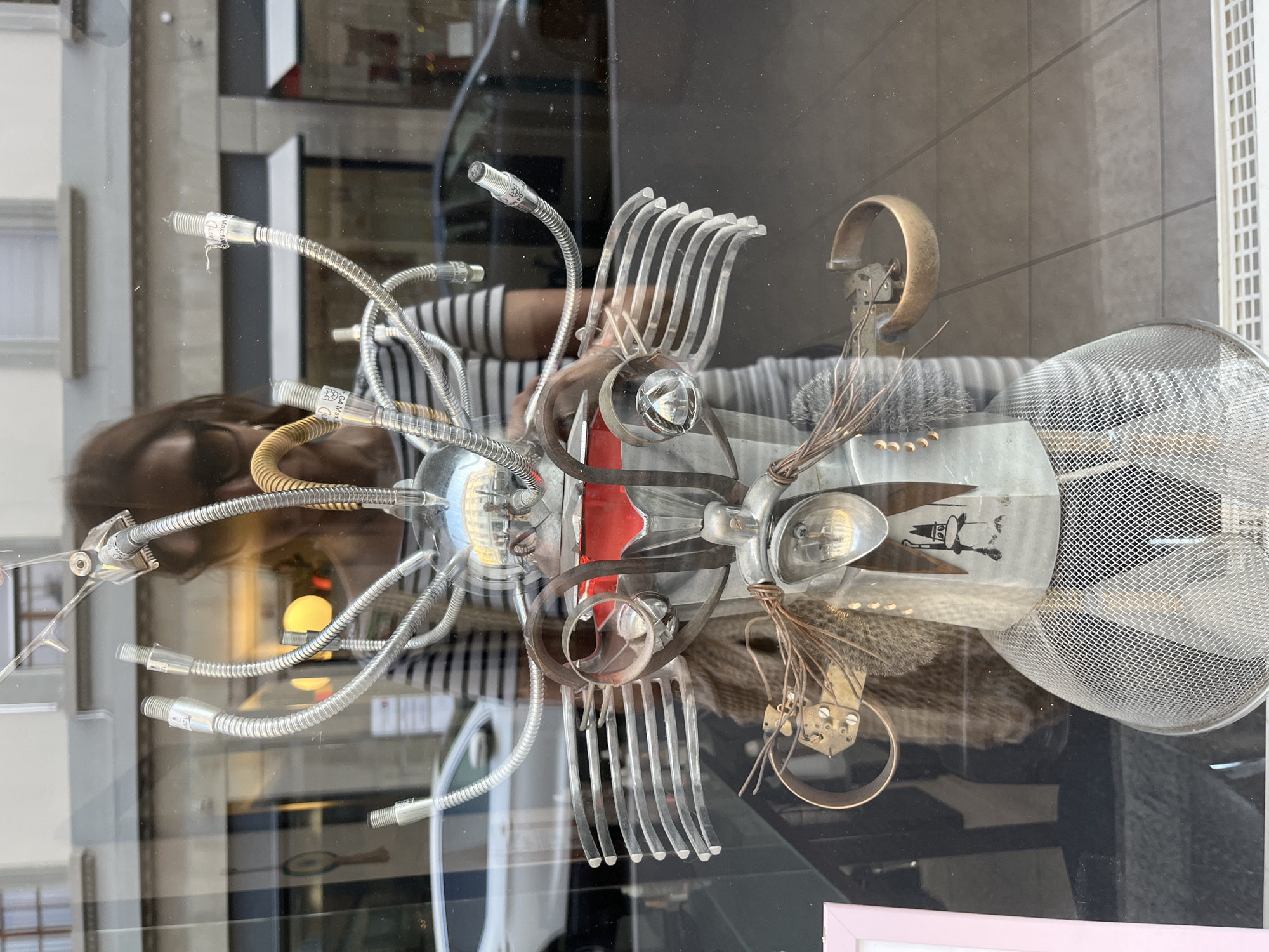 The image shows a whimsical, robotic sculpture displayed in a shop window in Vevey, Switzerland. The sculpture, made from various repurposed materials, stands prominently in the foreground. It features a metallic head with flexible tubes extending upwards, resembling antennae or hair. The body is composed of layered metallic elements, including what appear to be car grill parts and a red, jewel-like insert. There's a mesh-like lower body or skirt and decorative wire details.
Reflected in the glass of the shop window, a woman wearing a horizontally striped shirt and sunglasses is visible, holding a phone, presumably taking the photograph. The background shows a street scene with buildings, a white car, and a dark-colored car, suggesting an urban environment. The lighting indicates it might be late afternoon or early evening. The overall impression is a creative art installation viewed through a glass storefront.