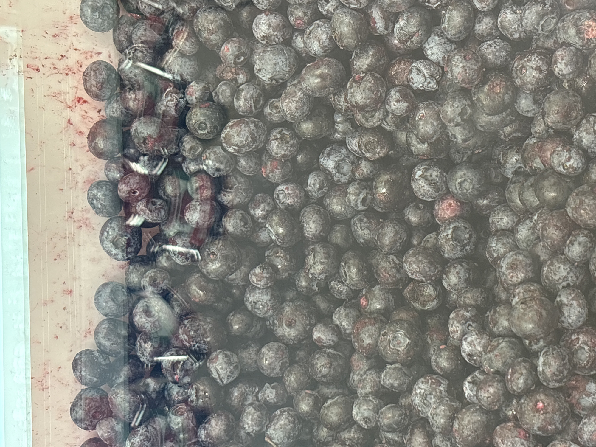 The image shows a close-up view of a container filled with blueberries. The berries are dark blue with a powdery coating, and some appear slightly crushed or bruised. The container is transparent, with a light-colored rim visible on the left side. There is no indication of the location being Minsk, Belarus, or any other specific place. No people, actions, or interactions are depicted in the image.