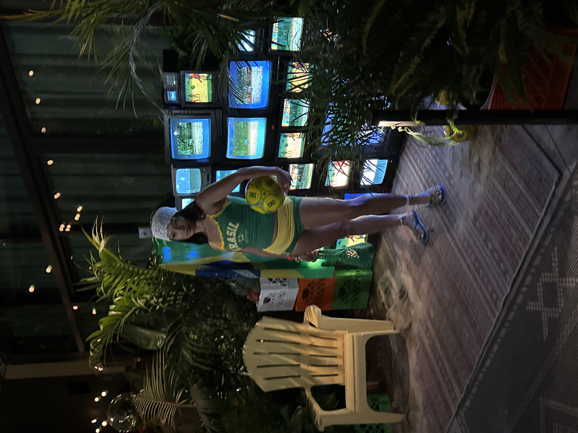 The image depicts an outdoor scene at night, possibly in Los Angeles, California, given the context of a tropical, beach-themed setup. A woman wearing a green and yellow outfit with a white bandana stands in the center, holding a soccer ball. Behind her is a wall of stacked vintage televisions, many displaying soccer-related imagery. To her left, a yellow plastic Adirondack chair sits on a patterned rug. The environment is enhanced with lush green foliage and palm plants, suggesting a recreational or event space. The lighting is dim, with some ambient lights visible in the background.