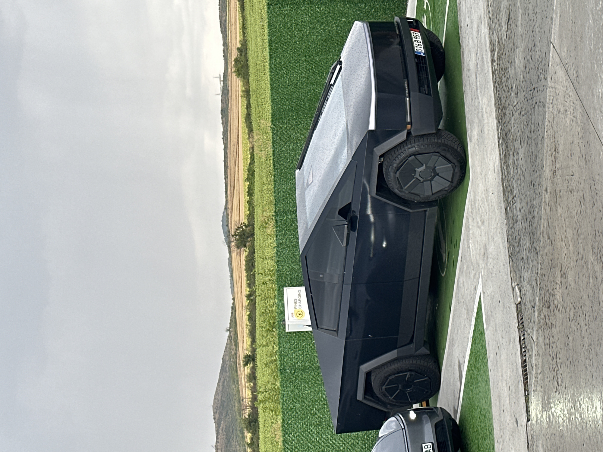 A black Tesla Cybertruck is parked at a charging station in Polsko Padarevo, Bulgaria. The truck is in the foreground, facing to the right. Its angular, stainless steel body is visible, as are its large, uniquely designed wheels. A charging sign with "FINES CHARGING" written on it is partially visible behind the truck.
In the background, there is a green hedge, followed by rolling hills and fields. The sky is overcast and gray, suggesting a cloudy day. Another vehicle, a dark-colored car, is partially visible to the left of the Cybertruck. The ground around the charging station is a mix of concrete and green artificial turf.