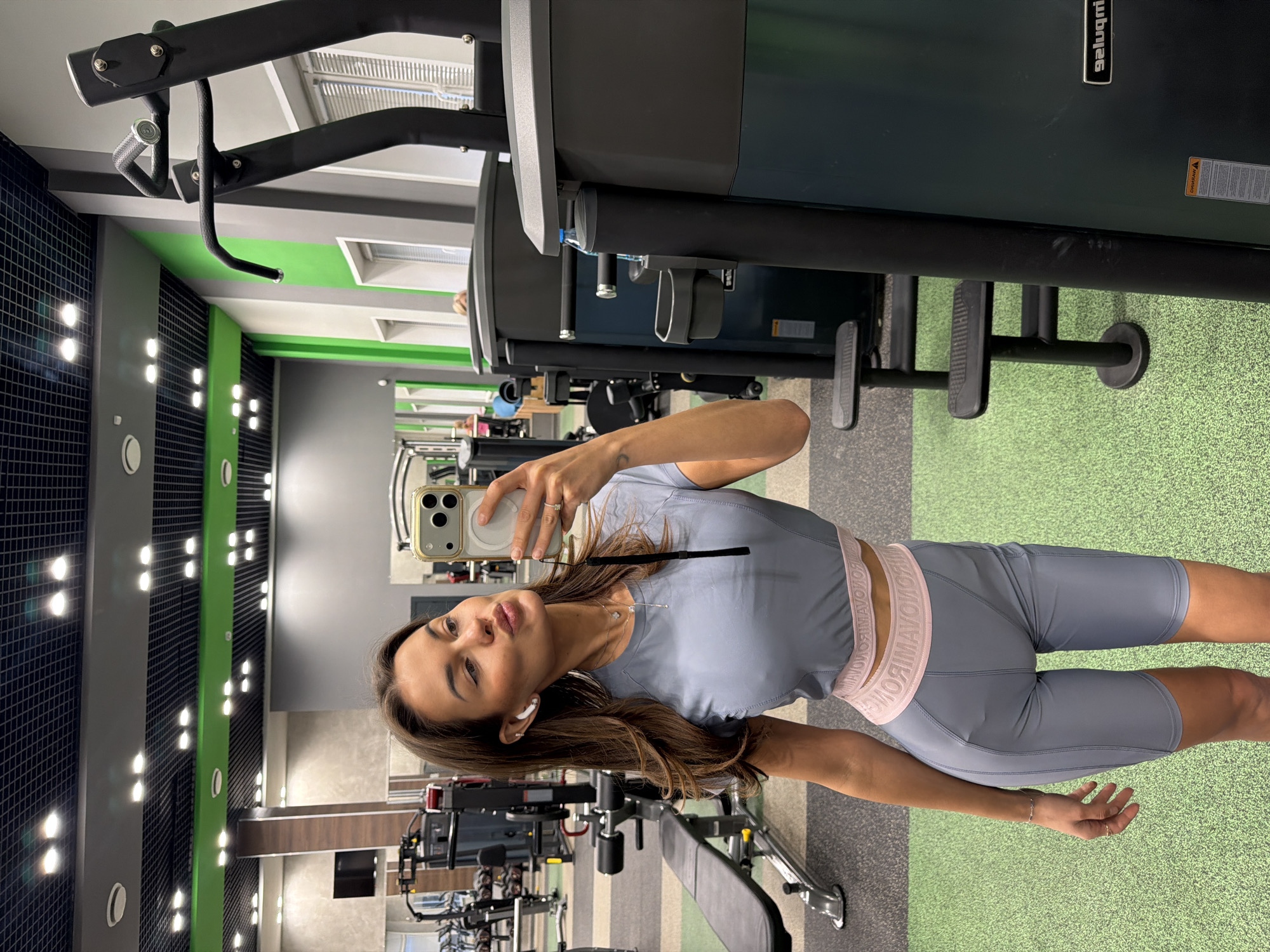 A woman is taking a selfie in a gym. She is wearing gray athletic shorts and a matching crop top with a pink waistband. She is holding a smartphone up to take the picture. The gym has various exercise machines visible, including weightlifting equipment and treadmills. The walls are gray and green, and the floor is covered with a green and gray rubberized surface. The gym appears to be located in Krasnoyarsk, Russia.
