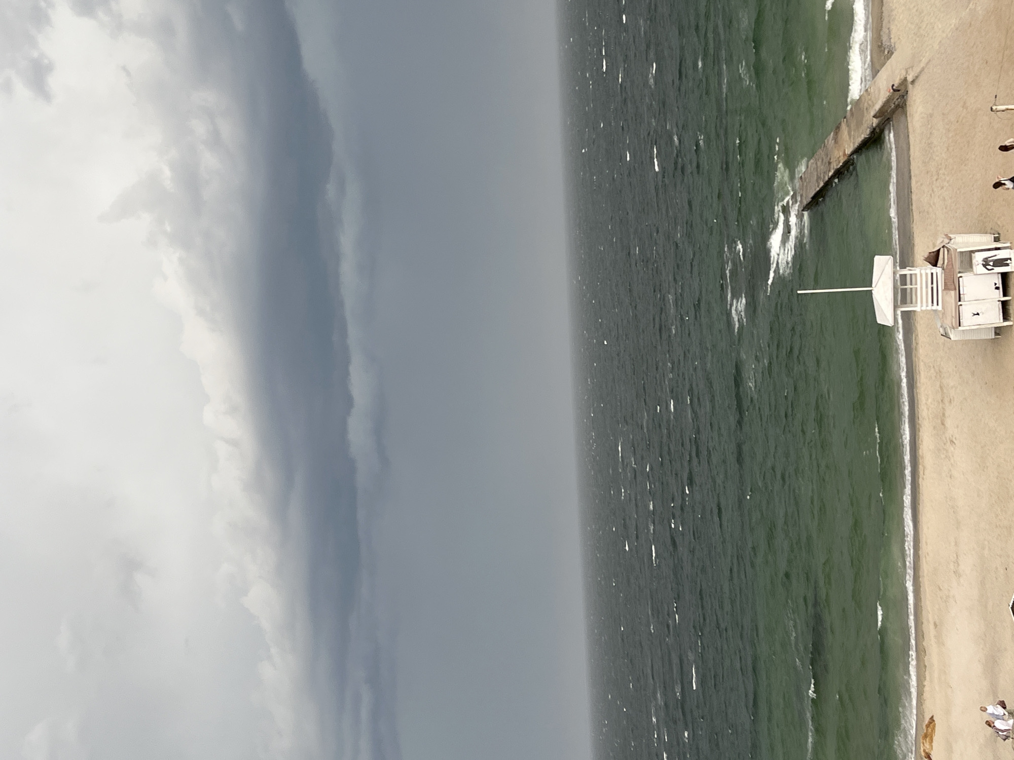 The image shows a beach scene, likely at the coast in the city of Unknown, Unknown. The sky is heavily overcast with dark storm clouds dominating the left side of the frame. The sea is a dark green color with some visible whitecaps and small waves. A concrete pier extends out into the water. The beach is sandy and contains a lifeguard station, constructed of white and off-white painted wood. There are a few people scattered on the beach near the lifeguard station.