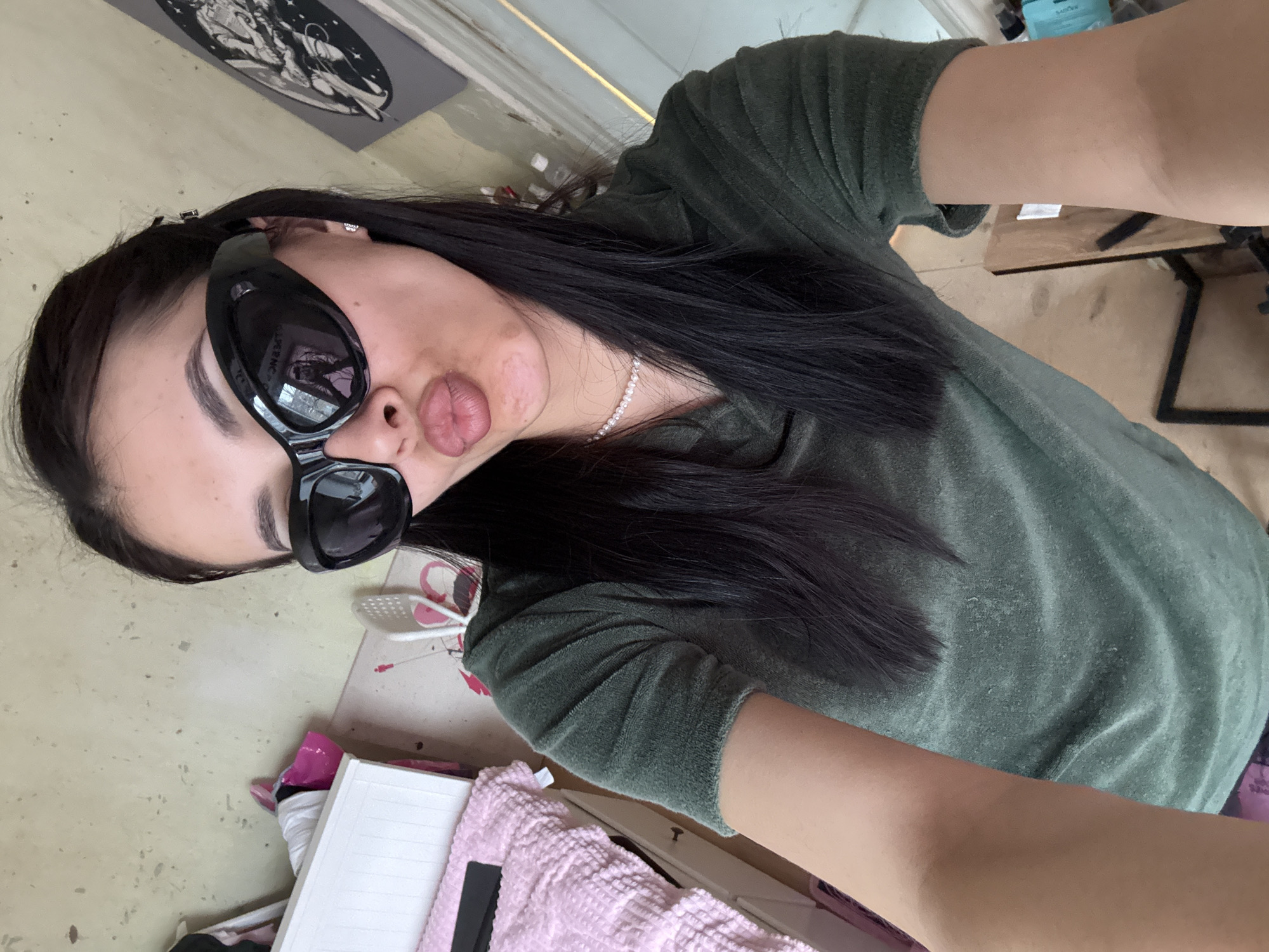 A medium shot captures a woman in a casual green t-shirt, wearing large, dark sunglasses and a pearl necklace. She is posed with pursed lips, suggesting a "kissing" face. Her dark hair falls across her shoulder.
The background reveals a cluttered indoor scene. A desk or table is visible, covered with various items including a pink blanket, a white notebook, and scattered objects. A framed illustration hangs on the wall, adding to the overall impression of a disorganized, personal space.
There is no information that indicates the location of the subject is in Votkinsk, Russia.