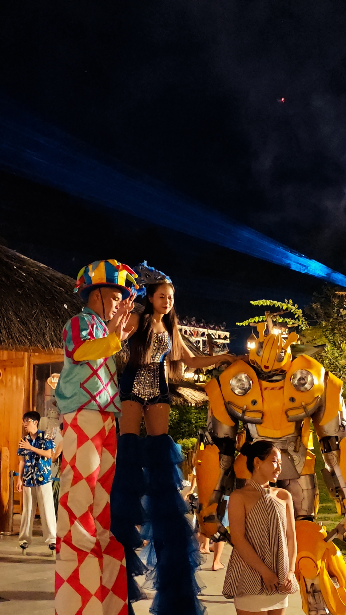 A nighttime outdoor event is captured in this photograph, possibly a festival or performance in Hoa Lư Ward, Vietnam. The scene is illuminated by blue spotlights and various ambient lights.
In the foreground, a person in elaborate clown-like stilts, dressed in a colorful costume with a jester's hat, stands with their hands raised as if in greeting or performance. Beside them, a young woman in a sequined blue outfit and tall blue ruffled legwear also stands on stilts, looking towards the right.
To their right, a large, bright yellow robot costume, resembling Bumblebee from Transformers, is prominently displayed. A woman in a striped sleeveless dress stands in front of the robot, looking to the side.
In the background, a young man in a blue floral shirt and light pants is visible on stilts, also appearing to be part of the performance. Other indistinct figures and structures, including a thatched roof building, are present in the background, suggesting a resort or entertainment venue. The night sky above is dark, with a faint red light visible in the upper right corner, possibly a distant light source or a drone.