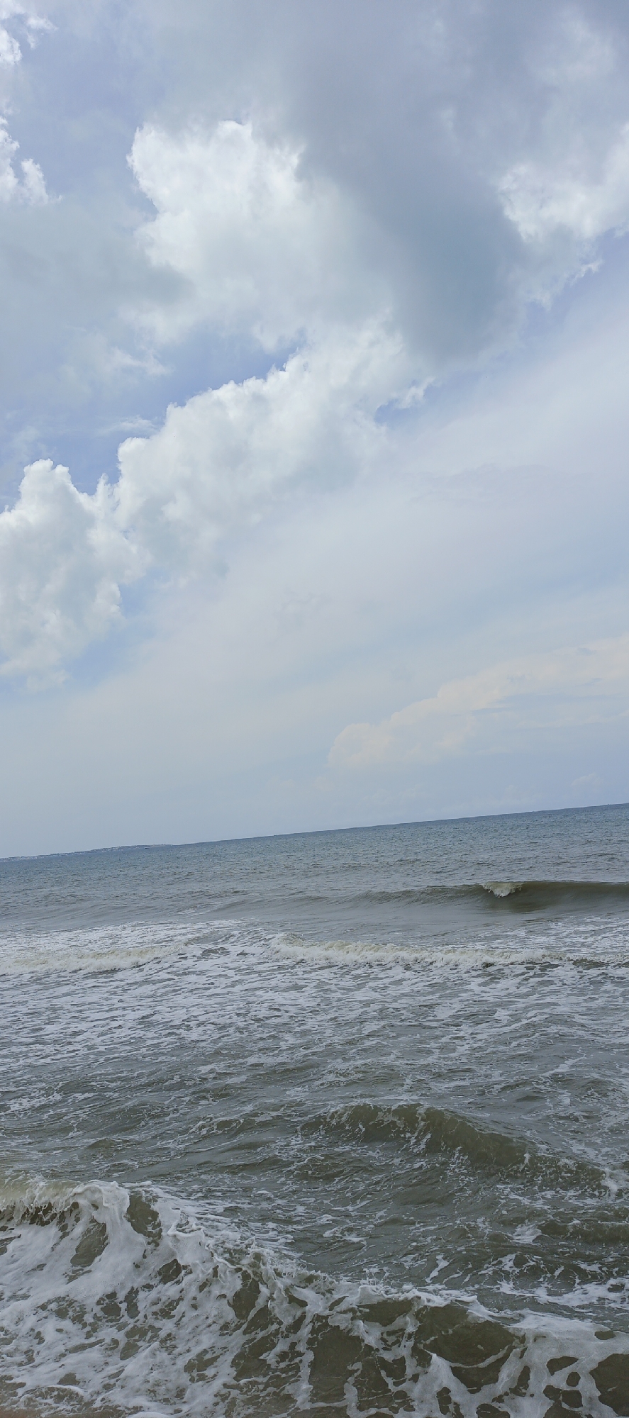 The image shows a wide shot of the ocean with a partially cloudy sky. The ocean is a deep blue-grey and appears to be in motion with visible waves. The waves in the foreground are breaking and creating white foam. The sky is a mix of light blue and grey clouds, with some clouds appearing fluffy and white. The horizon line is visible, separating the sea from the sky. The location is identified as Unknown, Unknown. There are no people or specific objects visible in the scene, only the natural elements of the ocean and sky.