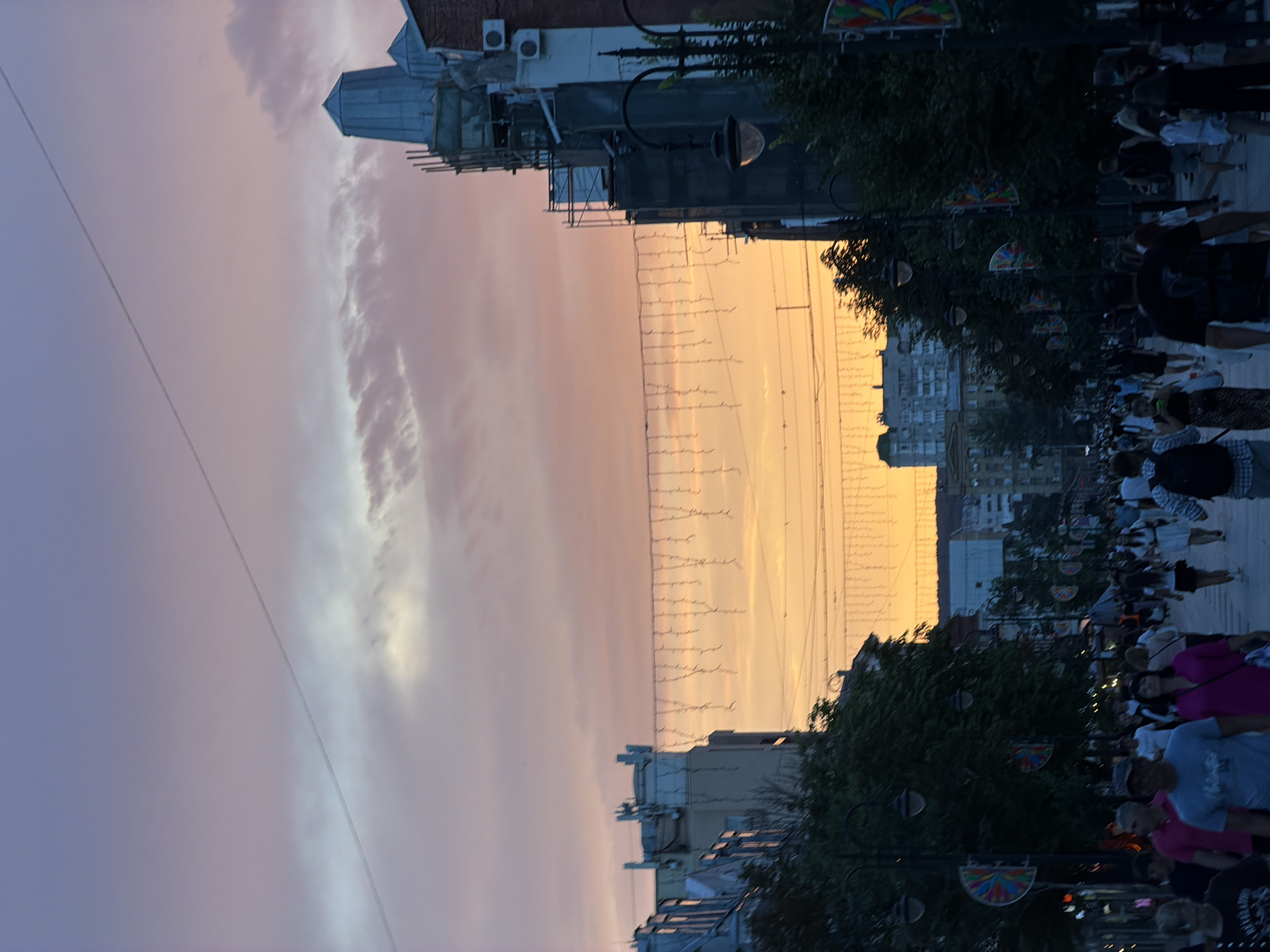 This is a vertical, eye-level, full-shot photograph taken at dusk on Prospekt Kirova in Saratov, Russia. The sky displays a gradient of pastel colors, transitioning from a pale blue at the top to hues of pink and orange closer to the horizon. A thin, dark power line bisects the sky from left to right.
Below the sky, the scene depicts a bustling pedestrian street lined with buildings on both sides. The buildings vary in architectural style and height, with some appearing older and ornate, while others are more modern and under construction, evidenced by scaffolding on one structure. Decorative string lights are hung across the street, adding a festive ambiance.
Numerous people are walking along the street, suggesting a lively evening atmosphere. They are captured in various poses, some walking alone, others in groups, and some appear to be shopping or socializing. The people are diverse in age and attire, wearing casual clothing appropriate for an evening stroll. Some individuals are walking towards the camera, while others are moving away.
Trees and streetlights are also visible along the sides of the street, with flags or banners attached to some of the lampposts. The overall impression is of a vibrant urban environment during a beautiful sunset.