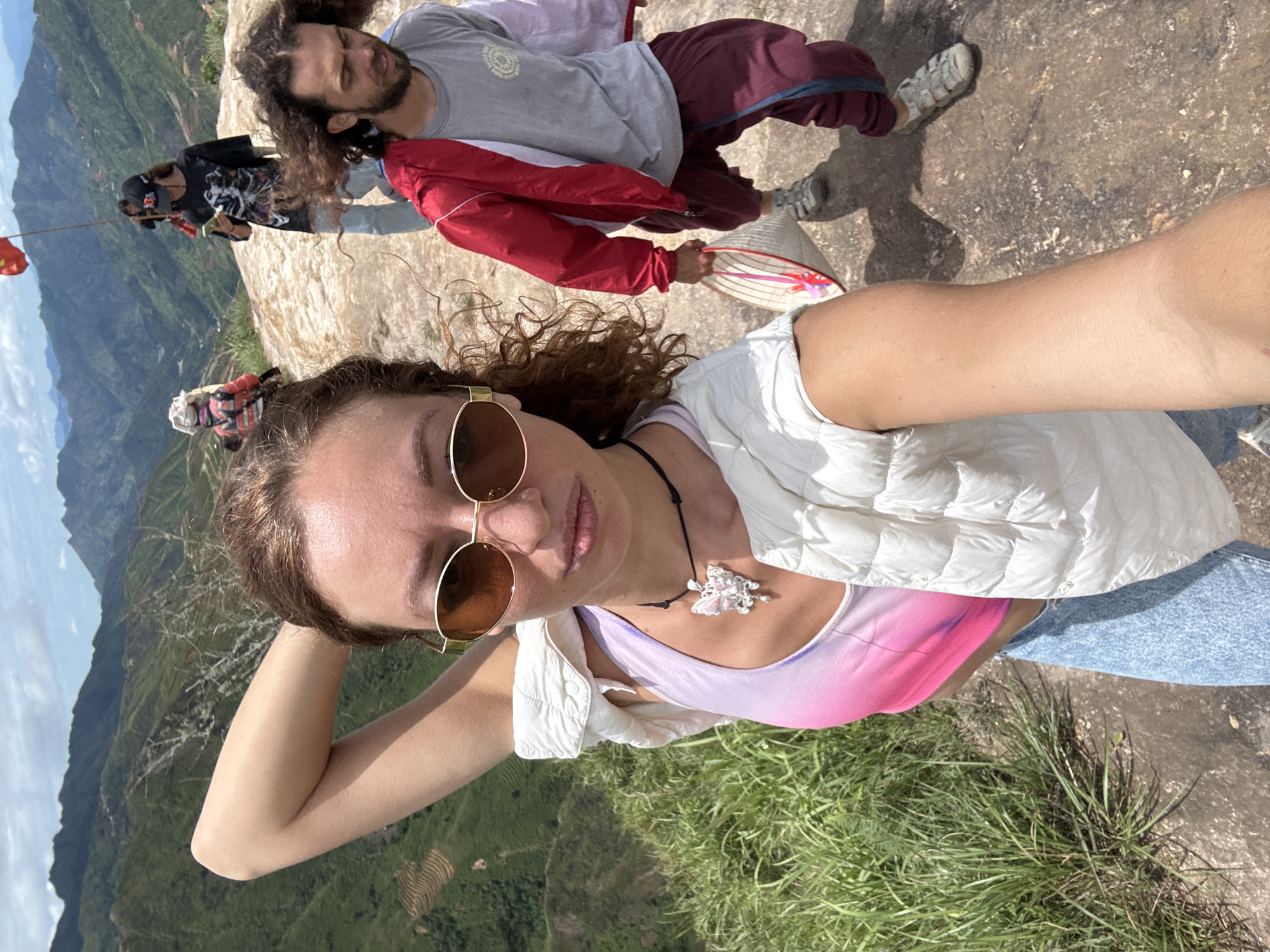 The image captures a group of people on a rocky, elevated viewpoint overlooking a lush, mountainous landscape in Ta Xua, Vietnam. The foreground features a woman in the center taking a selfie, her right arm raised behind her head, wearing a white puffer vest over a pink and purple tie-dye tank top, and light blue jeans. She sports oval, gold-rimmed sunglasses and a necklace with a pink butterfly pendant. To her right, a man with long, curly brown hair is walking away, wearing a red and white jacket over a grey t-shirt and maroon pants. He is carrying a conical straw hat. In the background, several other individuals are visible, including a person in a black t-shirt with a skull graphic and a baseball cap, looking at their phone, and another person in a bright, multi-colored jacket. A red flag on a pole stands in the background. The environment is characterized by steep, green, terraced mountains under a partly cloudy sky.