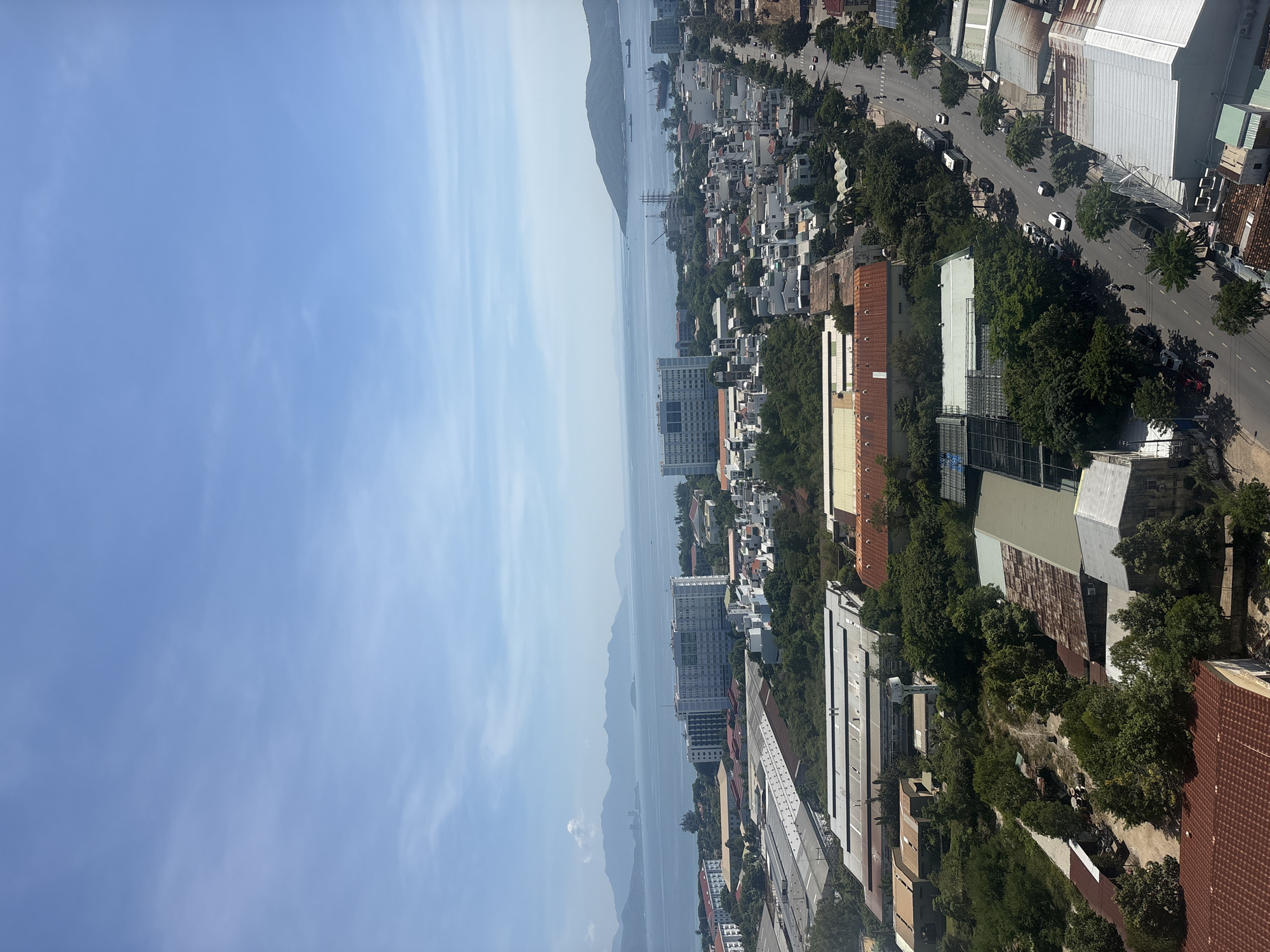This is an aerial, long shot photograph overlooking a coastal city in Nha Trang, Vietnam. The sky is a clear, light blue with a few wispy clouds. In the distance, a mountain range stretches across the horizon, bordering a calm, blue ocean. Several ships and boats are visible on the water.
The city below is a mix of residential buildings, industrial structures, and lush green trees. The residential area features white and light-colored buildings with red-tiled roofs, clustered together. The industrial section consists of large, flat-roofed warehouses with corrugated metal roofing. A multi-lane road runs through the city, with cars and motorcycles visible. People can be seen walking on the sidewalks and riding motorcycles.
The overall impression is a sunny day in a bustling coastal city, showcasing both its urban development and its proximity to natural landscapes.