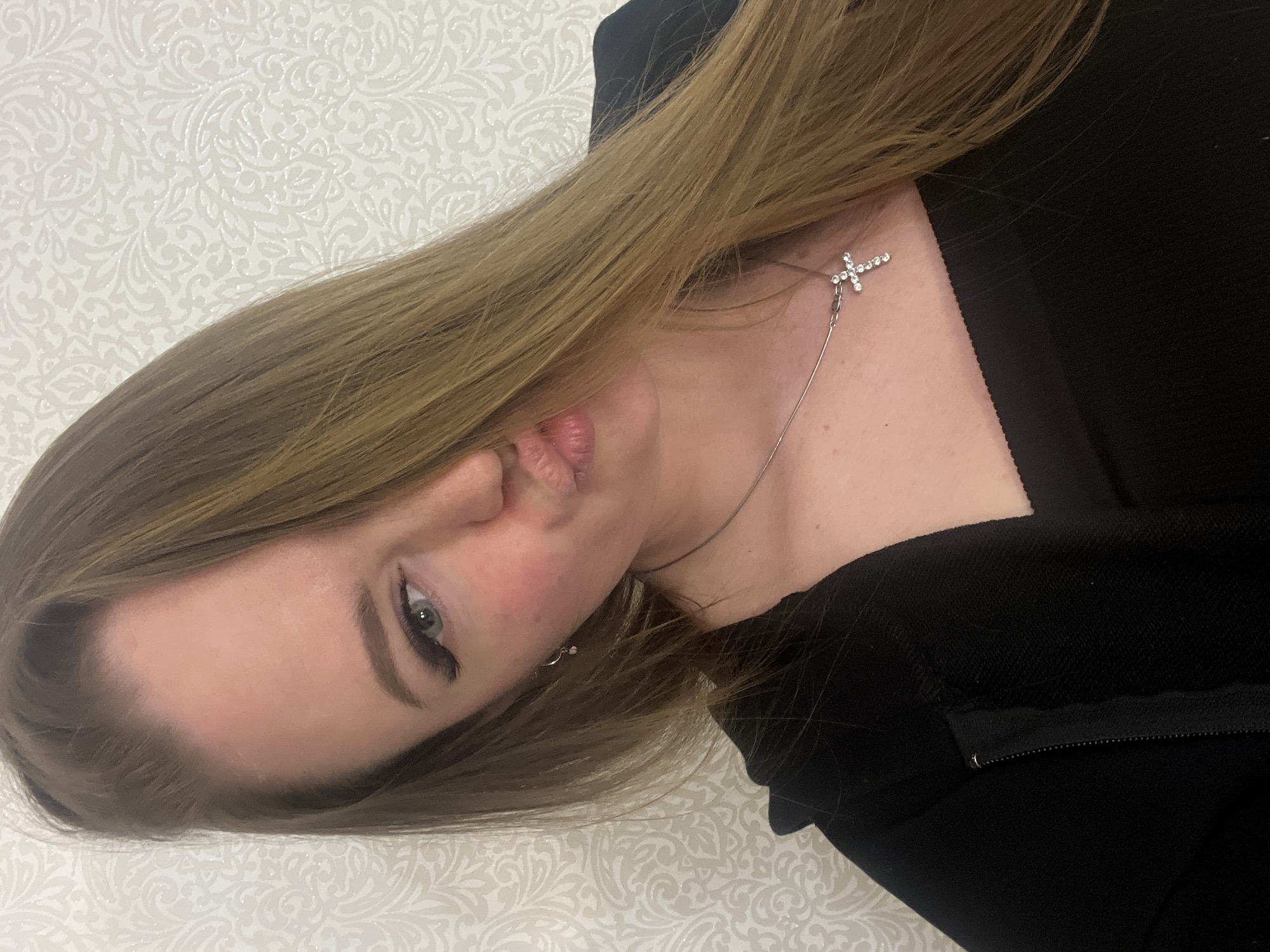 A close-up, portrait-style photograph captures a young woman posing indoors, identified as being in Sevastopol, Ukraine. Her head is tilted, and she looks directly at the camera with a slight pout or kiss face. She has long, light brown hair that flows down, partially covering her chest. Her eyes are light-colored, enhanced with prominent eyeliner and well-defined eyebrows.
The woman is wearing a silver-toned necklace featuring a sparkly, cross-shaped pendant. A small, silver hoop earring is visible in her left ear. Her attire appears to be dark, likely black, clothing, possibly a top or jacket, with a zipper detail visible on the right side of the frame.
The background consists of a light-colored wall decorated with a subtle, textured, ornate damask-like pattern. The lighting suggests an indoor setting. There is no visible text in the image. The overall tone of the photo is casual and personal.