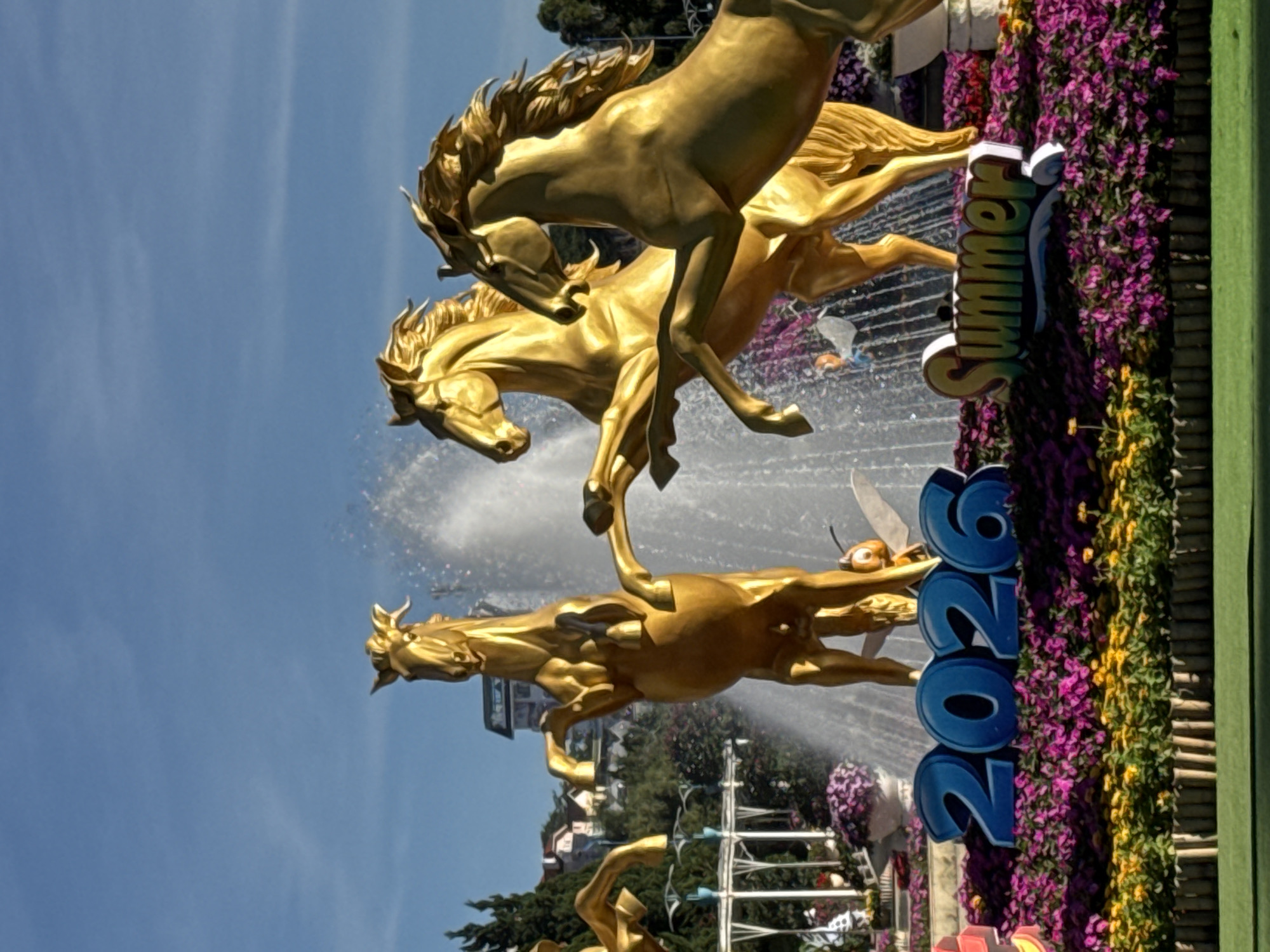 The image depicts a public garden or park decorated with golden horse sculptures, a fountain, and abundant floral arrangements. In the foreground, large blue numbers spell out "2026" and a colorful sign reads "Summer," both situated amidst vibrant purple and yellow flowers. Behind these, a fountain sprays water, partially obscuring three prominent golden horse sculptures, each dynamically posed. One horse stands on its hind legs, another rears up, and a third is positioned as if mid-gallop. A small, whimsical bee sculpture is also visible near the fountain. In the background, foliage and trees are present, along with buildings suggesting an urban setting. The sky is clear and blue. The scene is located in the Phường Xuân Hương area of Đà Lạt, Vietnam, a region known for its tourist attractions and floral displays.