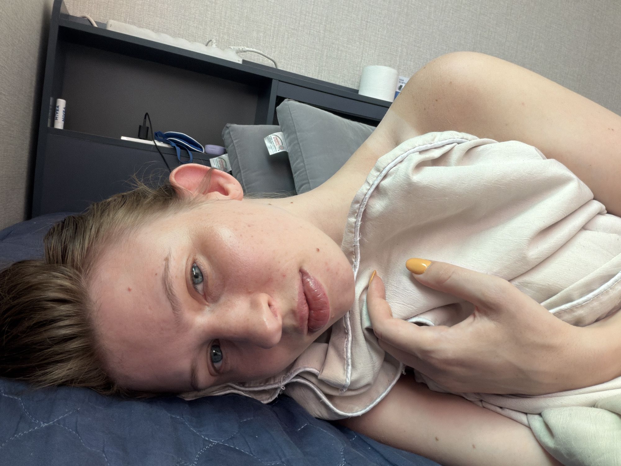 A young woman with light skin and blue eyes is lying on her side on a blue quilted surface. She has brown hair and is wearing a light-colored pajama top. Her right hand, with orange nail polish, rests on the fabric. In the background, there is a dark shelf with various items, including two Nivea products, a black charger, and a roll of toilet paper. Two grey pillows are also visible. The overall environment appears to be a bedroom, possibly in a residence in Dongducheon-si, South Korea.
