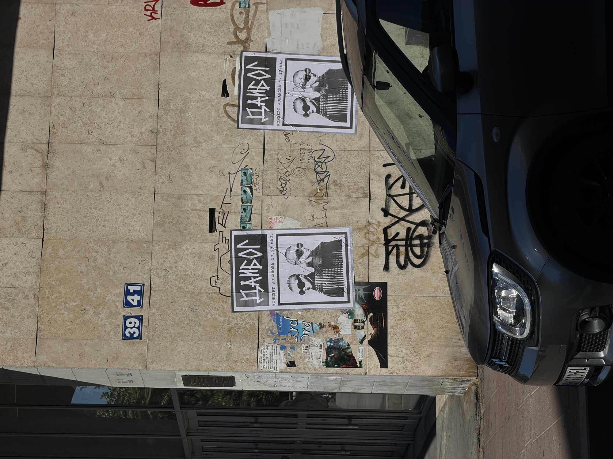 The image shows an exterior wall of a building in Belgrade, Serbia. The wall is made of light-colored stone tiles and is partially covered with graffiti. There are two posters advertising a concert by Даибол (DAIBOL) at Јованова 57 on May 27th. Below these posters are several other smaller posters and flyers. Blue address number plaques with the numbers 39 and 41 are visible on the left side of the wall. A dark grey car is parked next to the wall, partially obscuring the right side of the image. The front of the car, including the headlight and grill, are visible. The bottom of the image shows a modern glass and metal door and window. The overall scene suggests an urban environment with some decay and layers of urban culture.