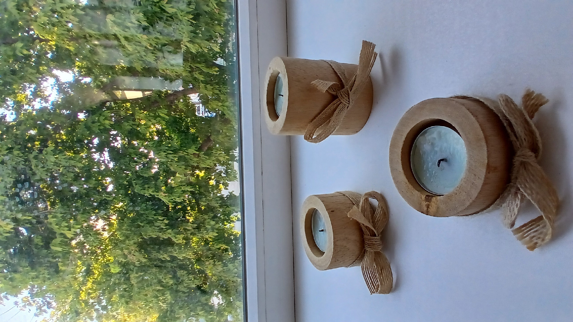 Three wooden tea light candle holders with twine bows are placed on a white surface in front of a windowsill. Through the window, a dense green tree with sunlight filtering through its leaves is visible. The scene is lit by natural light. The specific location within the city of Oral, Kazakhstan, cannot be determined from the image.