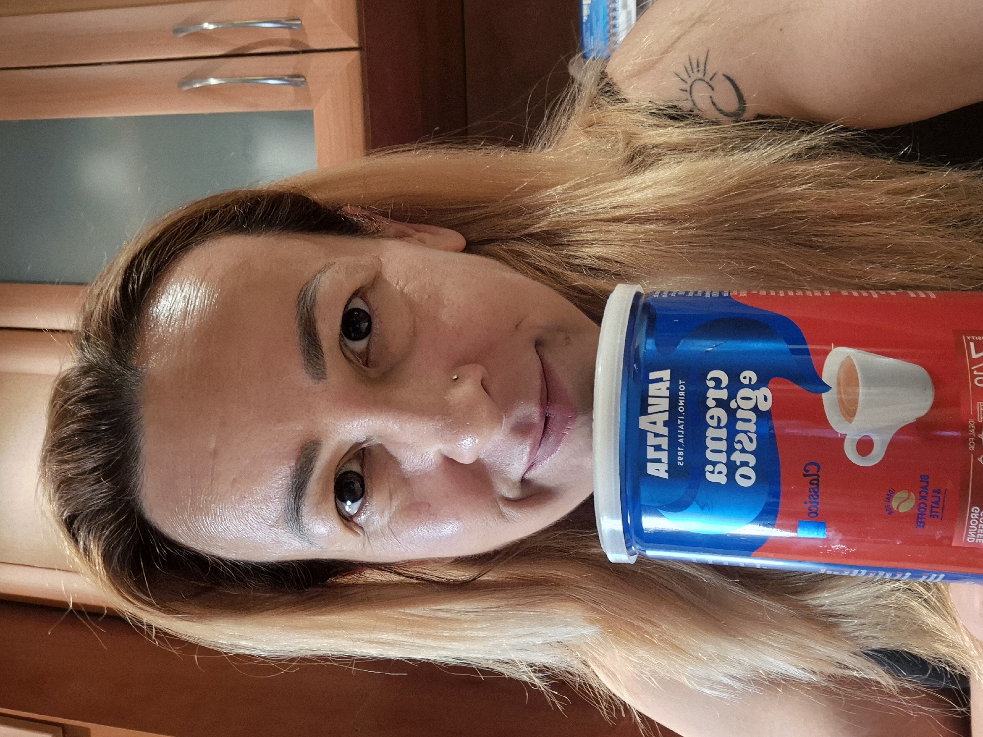A medium shot shows a woman with blonde hair and a nose ring holding a blue and red can of Lavazza coffee in front of her chest. She is looking directly at the camera with a slight smile. Her hair is long and wavy, falling to her shoulders. She is wearing a black sleeveless top and has a tattoo of a sun and crescent moon on her right shoulder. The background appears to be a kitchen with wooden cabinets. The woman's face is well-lit, and the details of her skin are visible, including some light wrinkles around her eyes and forehead. The can of coffee is the main object in focus besides the woman. The text on the can is partially obscured and some of it is upside down and mirrored, indicating that the can is being held at an angle or has been rotated. The setting is indoors, in what appears to be a domestic environment.
