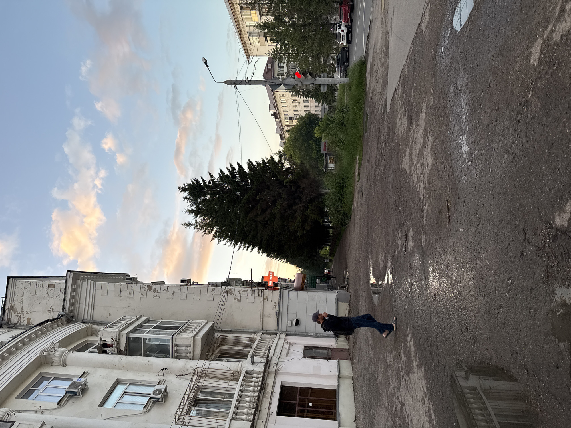 A man in a dark hoodie and jeans walks on a wet asphalt sidewalk next to a white building. The building has multiple windows and balconies with ornate railings. A red pharmacy sign with white lettering is visible on the side of the building. The sidewalk is dotted with puddles reflecting the sky and buildings. A line of tall evergreen trees borders the sidewalk. In the background, apartment buildings and a traffic light can be seen. The sky is a mix of blue and pink clouds, indicating either sunrise or sunset. The scene appears to be in an urban environment, potentially in Kazan, Russia, given the architectural style and the Cyrillic lettering on the pharmacy sign.